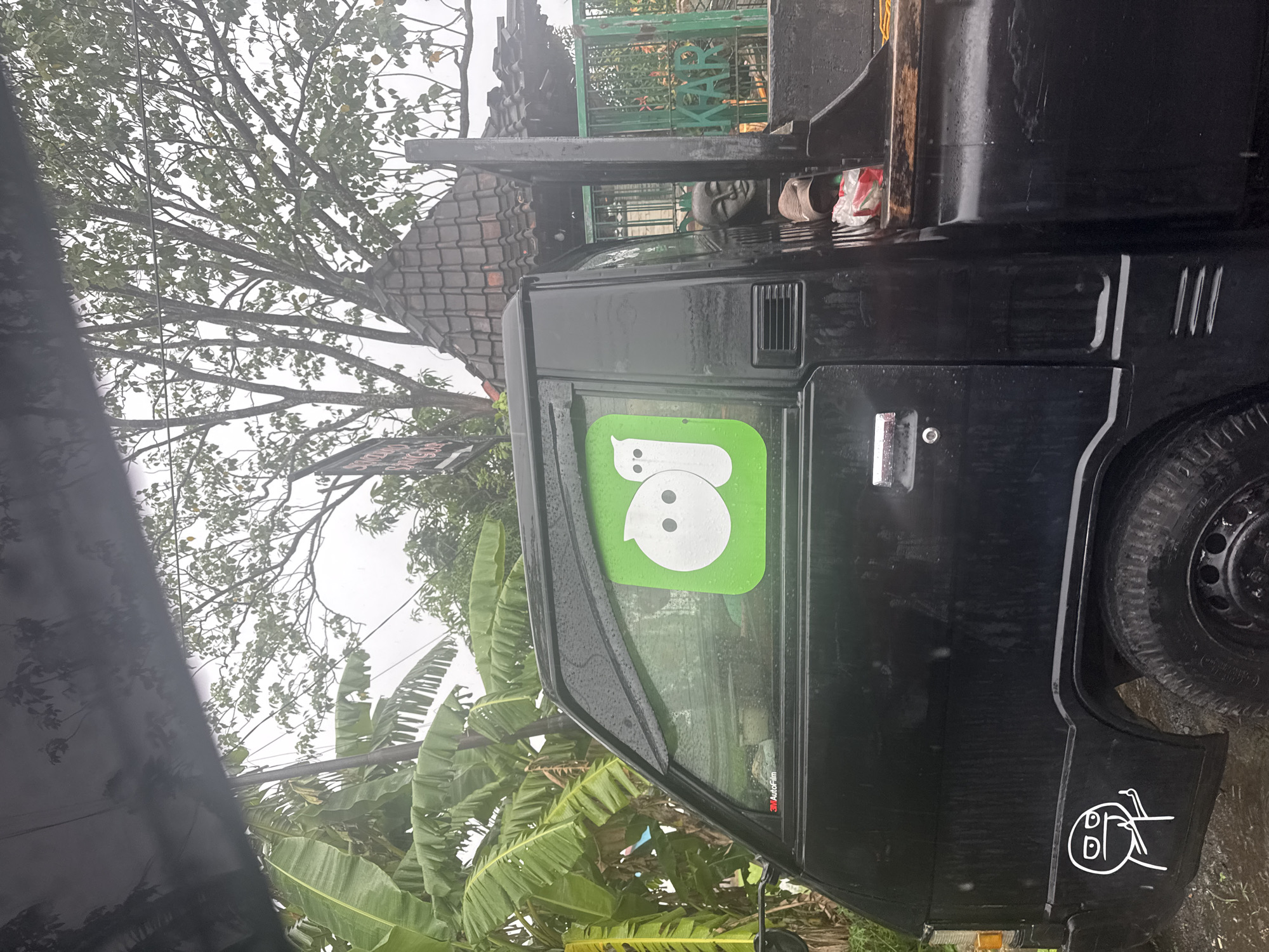 The image shows the side of a black vehicle in North Kuta, Indonesia. The vehicle has a green sticker with a white ghost figure on the window. A white sticker depicting a stick figure is located near the rear wheel. A tree and banana plants are visible in the background. A tiled roof is partially visible above the vehicle. The image was likely taken in wet weather as the vehicle surface appears damp.