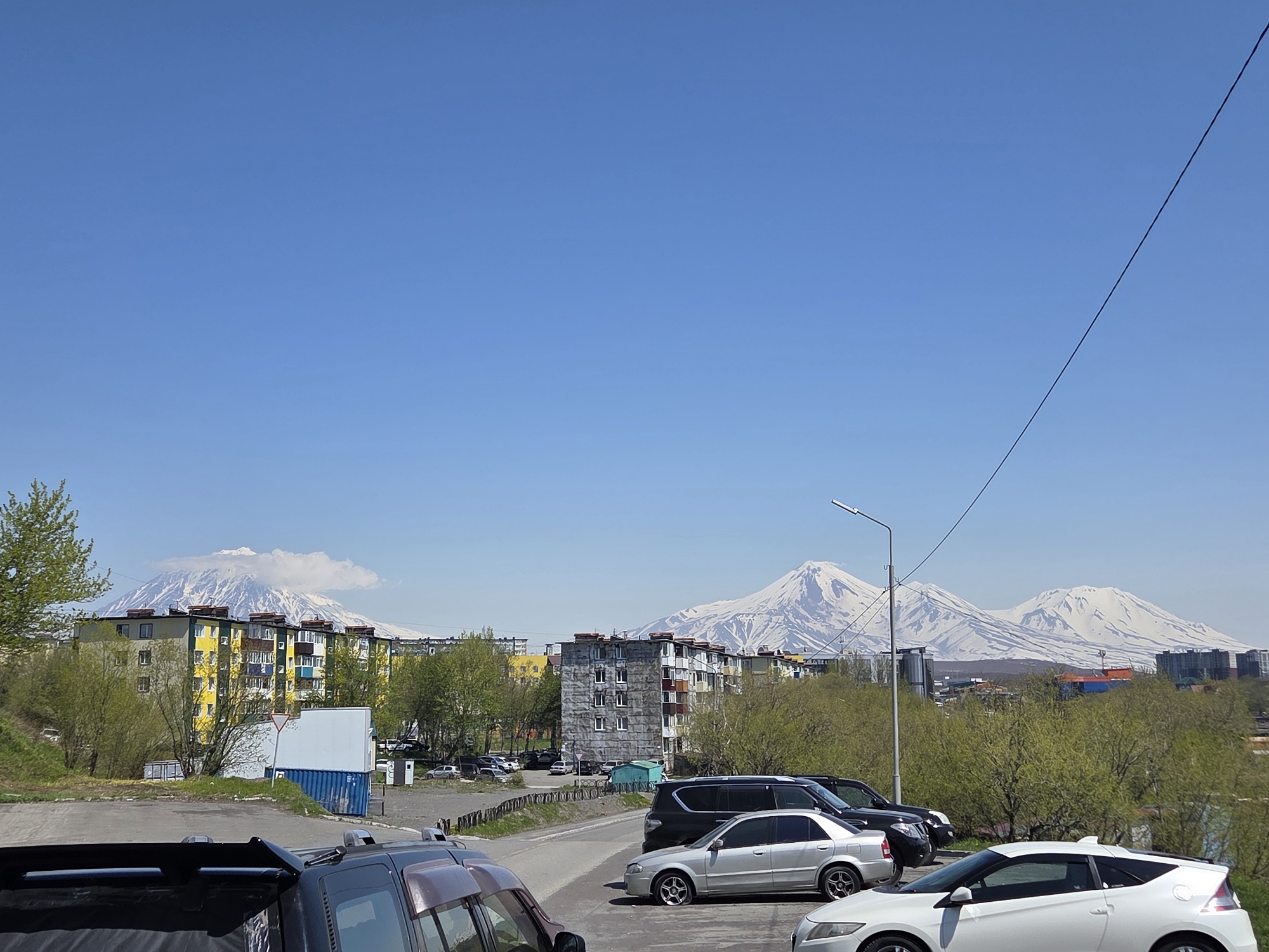 The image presents a panoramic view of Petropavlovsk-Kamchatsky, Russia, under a clear, bright blue sky. In the distant background, a series of snow-capped mountains, consistent with volcanoes, extend across the horizon. On the far left, one large peak is partially enveloped by a distinctive cloud formation near its summit. To the right, a prominent cluster of three snow-covered volcanoes, identifiable as Koryaksky, Avachinsky, and Kozelsky, rises distinctly.
The mid-ground features urban elements, including several multi-story residential apartment buildings. On the left, a building with yellow and blue facades stands out, adjacent to a more uniformly gray structure. The landscape is interspersed with lush green trees and bushes, suggesting a spring or early summer season. A paved area or road runs through the mid-ground, leading to additional parked vehicles. A white rectangular structure, a red-trimmed "give way" sign, and a blue shipping container are visible in this section.
The foreground is dominated by parked vehicles and a paved surface. On the lower left, the rear section of a dark SUV or truck is visible. In the lower right, a white Honda CR-Z coupe is prominently parked, angled slightly to the left. Further back on the right, a silver sedan is parked alongside a dark SUV or van. A street lamp with an overhead power line diagonally extends from the upper right toward the center. The entire scene is illuminated by strong natural sunlight. No human figures are present.