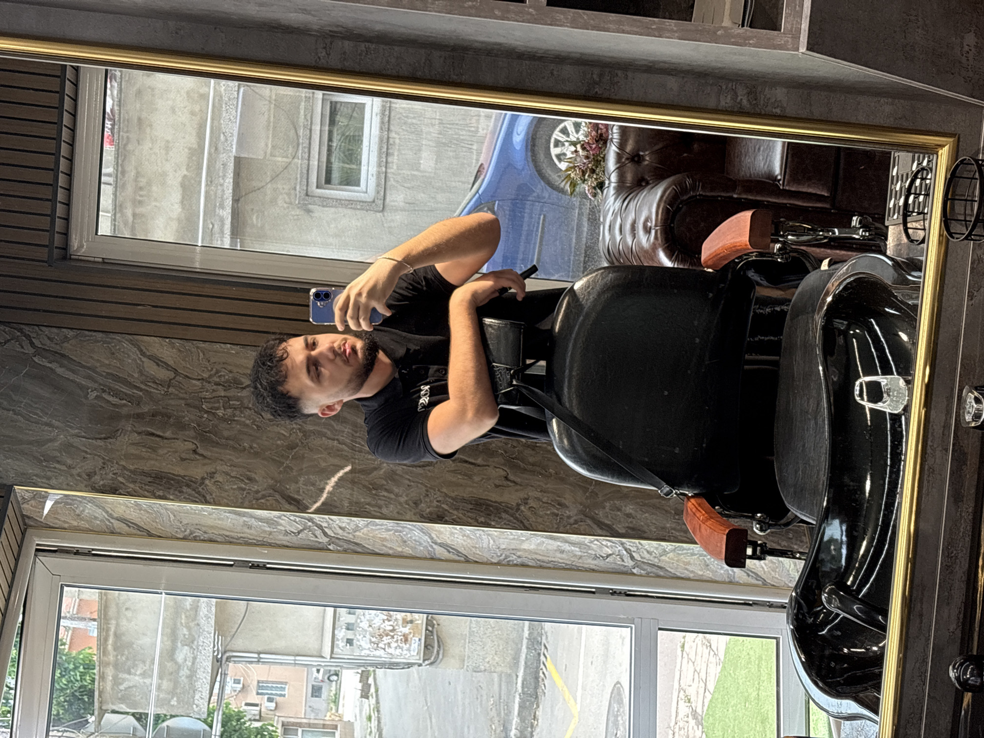 A young man with short, dark curly hair and a beard is captured taking a selfie in a large mirror within what appears to be a modern barber shop or salon. He wears a black short-sleeved t-shirt, on which the word "DREAM" is partially visible. He holds a blue smartphone in his right hand, pointing it towards his reflection. His left arm rests on the armrest of a sleek, black barber chair with wooden accents.
The interior of the establishment features a prominent rectangular mirror with a golden frame, reflecting parts of the room. A large, dark brown tufted leather armchair or sofa is visible in the mirror's reflection, suggesting a comfortable waiting area. The walls are decorated with dark, marble-like panels and horizontal wooden slats, contributing to an upscale aesthetic.
Beyond the mirror and through a large window, an urban outdoor scene is visible under natural daylight, indicating it is daytime. This includes parts of a street with yellow markings and a manhole cover, various buildings made of concrete or stone, a blue car parked, and some green foliage. The setting suggests an urban location, consistent with Varna, Bulgaria. The man's expression is neutral, focused on capturing the image.