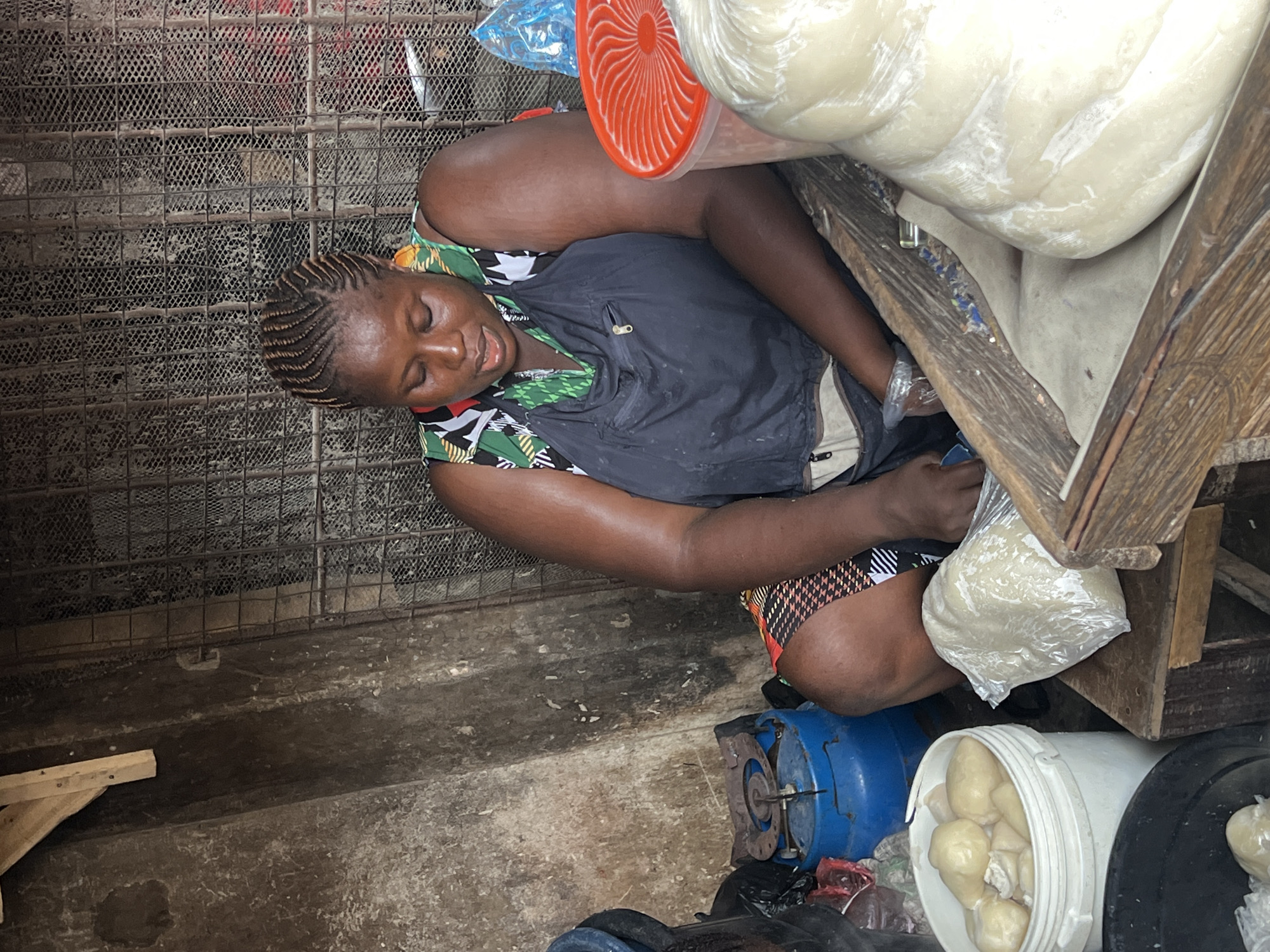 The image depicts a woman with her eyes closed, appearing to be resting or sleeping, in a market setting in Nkpor, Nigeria. She is seated next to a large, pale, dough-like substance wrapped in plastic, which is resting on a wooden structure. The woman is wearing a dark vest over a patterned shirt and shorts. Her hair is styled in neat braids.
In front of her, a blue gas cylinder is visible, along with a white bucket filled with similarly wrapped dough or food items. The ground appears to be concrete, with some visible debris. To the left of the woman, a metal grid or fence is present. The lighting suggests it could be daytime, but the specific time or weather conditions are not discernible. The overall impression is one of a busy, possibly humid, outdoor market environment. There are no visible text elements in the image.