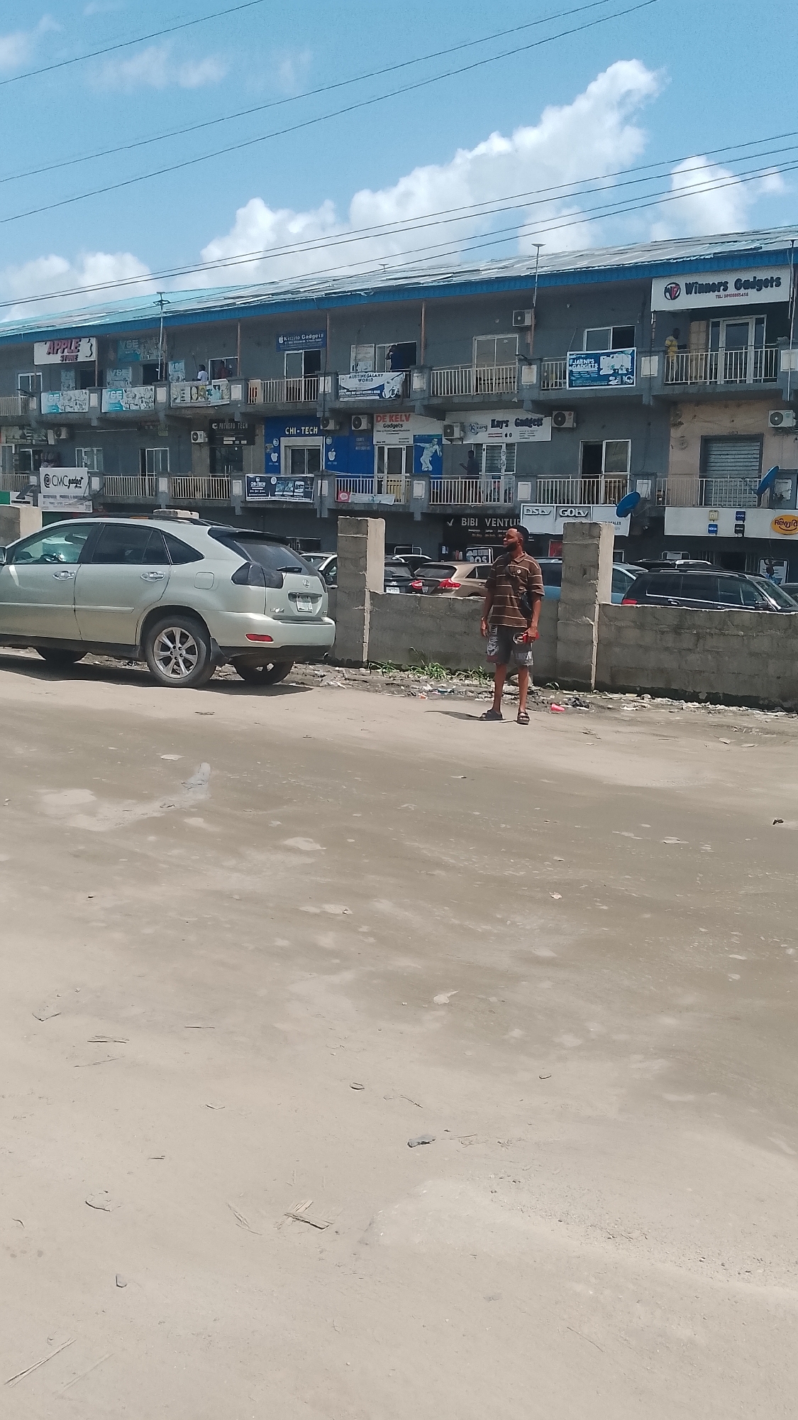 The image depicts a street scene in Effurun, Nigeria. A man stands near a low concrete barrier separating the street from a two-story commercial building. The building houses various businesses, indicated by signs such as "Winners Gadgets," "Kiszito Gadgets," "DE KELV Gadgets," "Lay's Gadgets," "CHI-TECH," "BIBI VENTURES," "CMC gadgets," and "APPLE store" among others. There are several cars parked behind the barrier. The road is dirt.