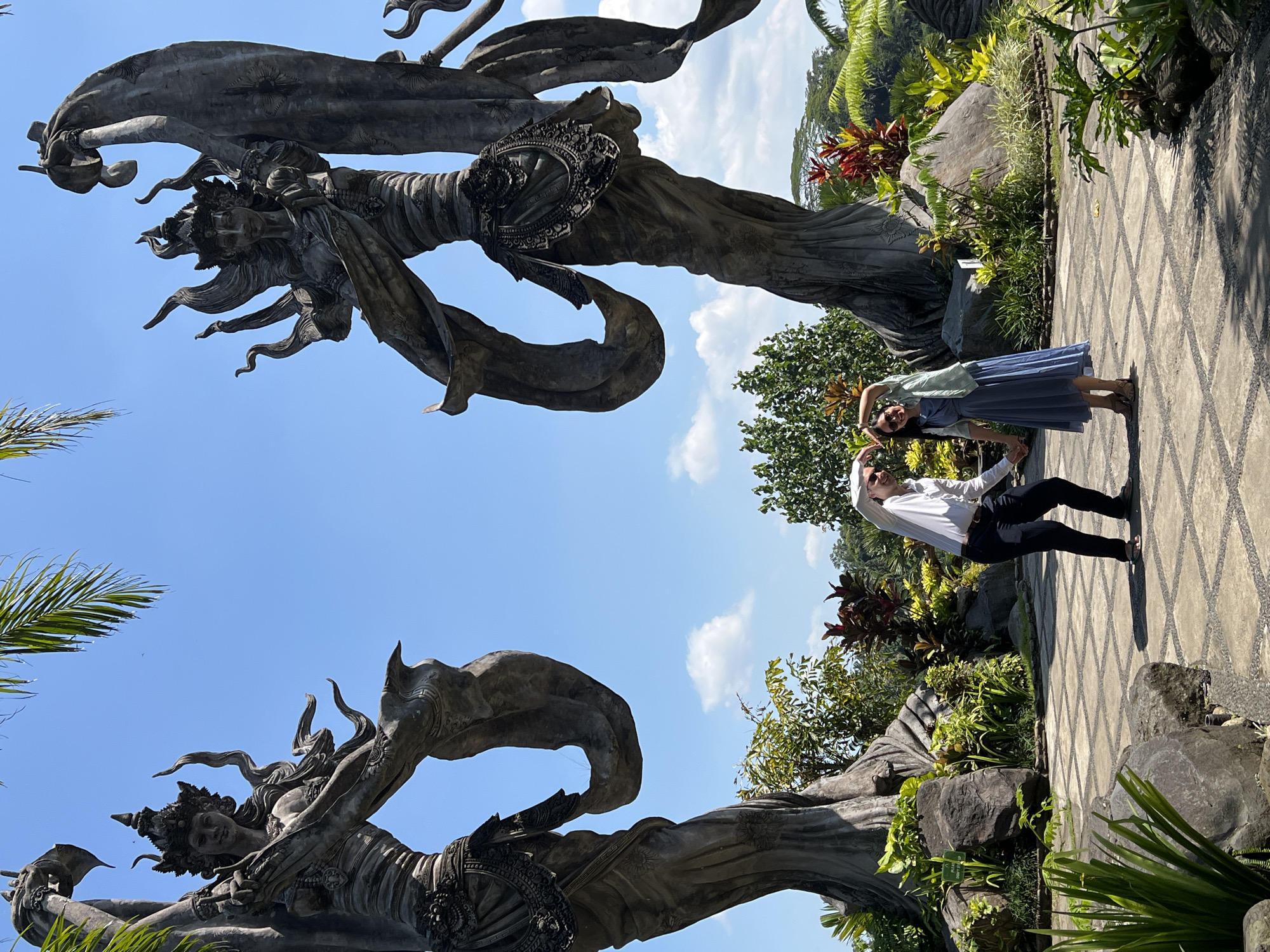 A couple is posing on a stone-tiled path in Kedewatan, Indonesia. The woman, on the right, wears a blue dress and a light-colored wrap. The man, to her left, wears a white shirt and dark pants. They are holding hands and forming a heart shape with their arms above their heads.
Two large, weathered stone sculptures flank the couple. The sculptures depict ornate, mythical figures with flowing drapery. Lush greenery, including trees, shrubs, and rocks, surrounds the path and sculptures. The sky is blue with scattered clouds.