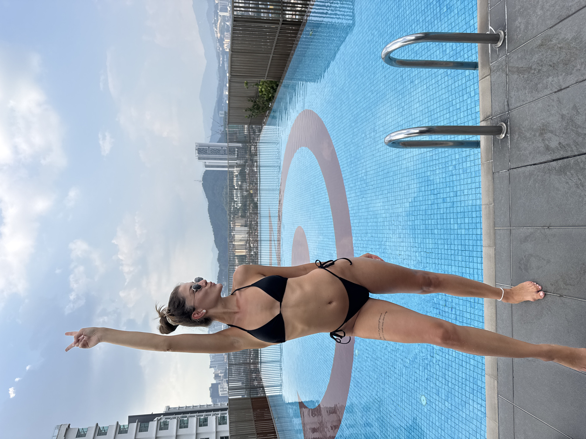 A woman in a black two-piece bikini poses beside a rooftop swimming pool, with a city skyline and mountains in the background under a partly cloudy sky in Kuala Lumpur, Malaysia. She is standing upright on the dark grey tiled poolside, leaning slightly back with her head tilted upwards, looking towards the sky. Her right arm is extended straight, pointing towards the upper left portion of the frame. Her left arm is bent at the elbow. She is wearing sunglasses, a single pearl stud earring in her right ear, and a white anklet on her right ankle. A tattoo with unreadable script is visible on her left thigh. The pool has bright blue mosaic tiles, with a large, light brown circular pattern on its floor. Two metal pool ladder handles are visible on the far side of the pool. The time of day appears to be bright daytime.