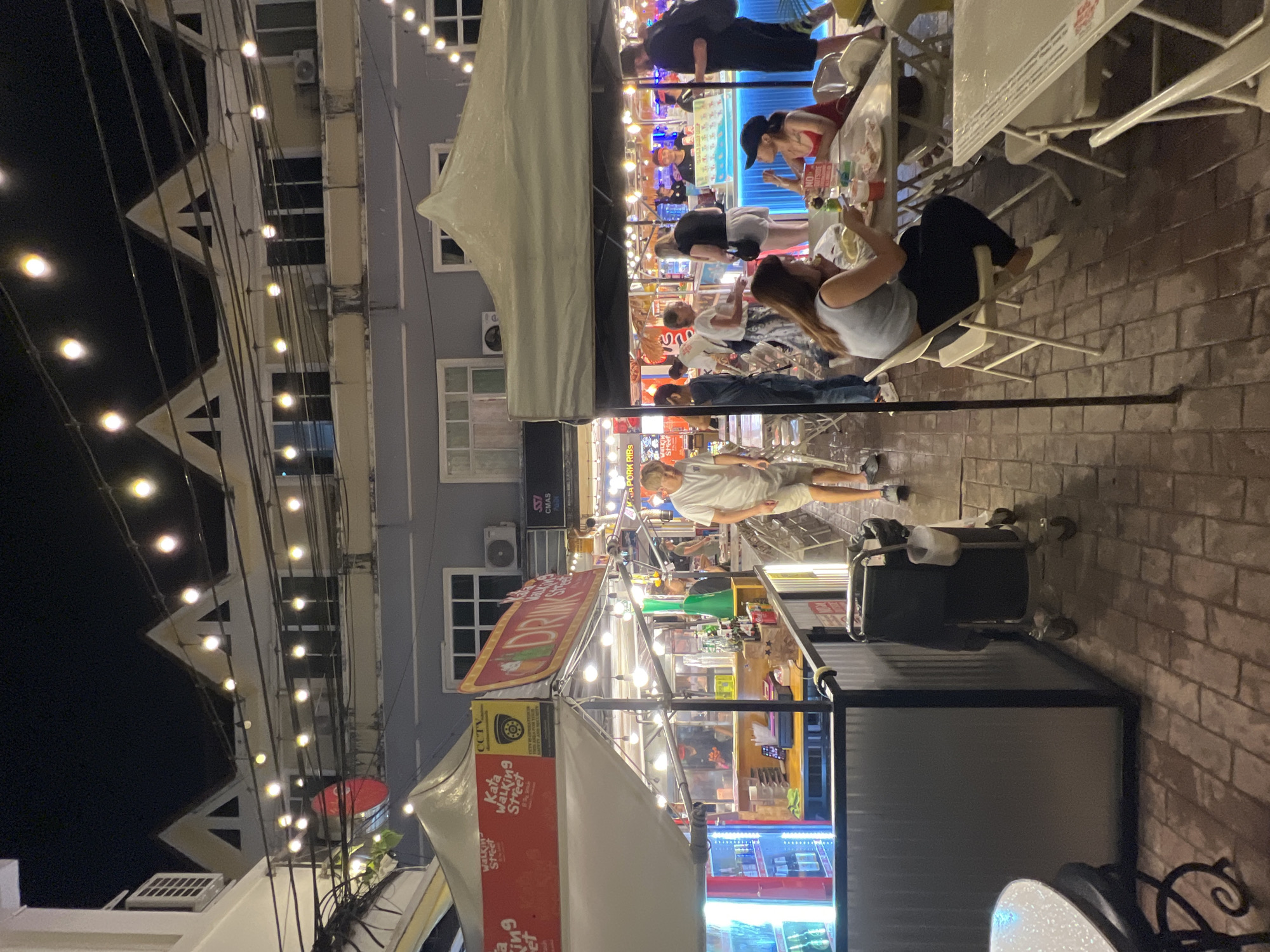The image depicts a lively night scene in Karon, Thailand, likely a bustling street food market or a vibrant entertainment area. The foreground is dominated by a white tent structure, possibly a stall or seating area, with prominent signage indicating "Kata Walking Street" and "DRINK". A red and yellow sign with "CCTV" is also visible.
Further into the scene, several people are gathered around tables and food stalls. One person, a man wearing a white t-shirt and light-colored shorts, is standing near a stall, appearing to observe the surroundings. Other individuals are seated, engaged in conversations or dining, creating an atmosphere of social interaction and enjoyment. The stalls are illuminated by strings of warm, bright lights, casting a festive glow on the brick-paved walkway and the buildings in the background.
The overall impression is one of a vibrant evening at a popular tourist spot. The presence of food stalls, people dining, and festive lighting suggests a lively atmosphere characteristic of a night market or a busy street in a tourist destination. The time of day is clearly nighttime, and the weather appears clear, with no indications of rain or adverse conditions. The buildings in the background, with their balconies and windows, suggest a multi-story structure adjacent to the market area. The activity taking place is people enjoying food and socializing in a lively night market setting. Visible text includes "Kata Walking Street," "DRINK," "CCTV," and "PORK RIBS."