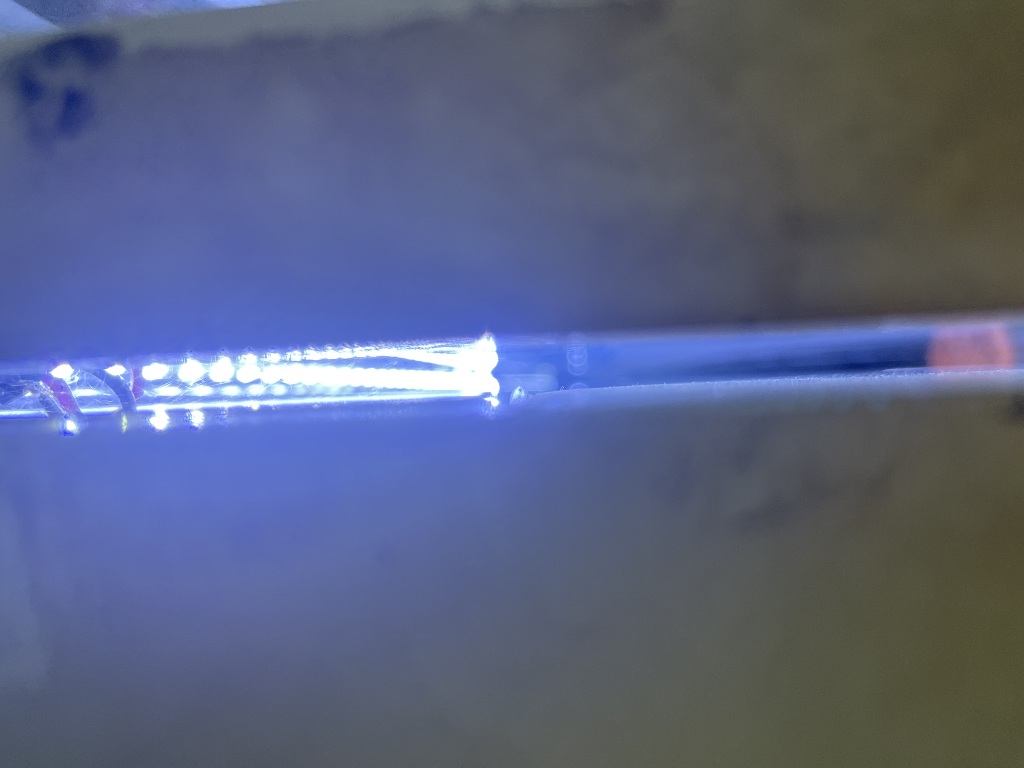 The image shows a close-up of a blue LED light strip against a blurred, neutral background. The LEDs are small and arranged in a linear fashion. The background appears to be a wall or a similar flat surface, with some subtle variations in color and texture, possibly indicating a dimly lit indoor environment. No specific location in Central Jakarta, Indonesia, can be identified from the visual information provided. The image is abstract and focuses on the light source and its immediate surroundings.