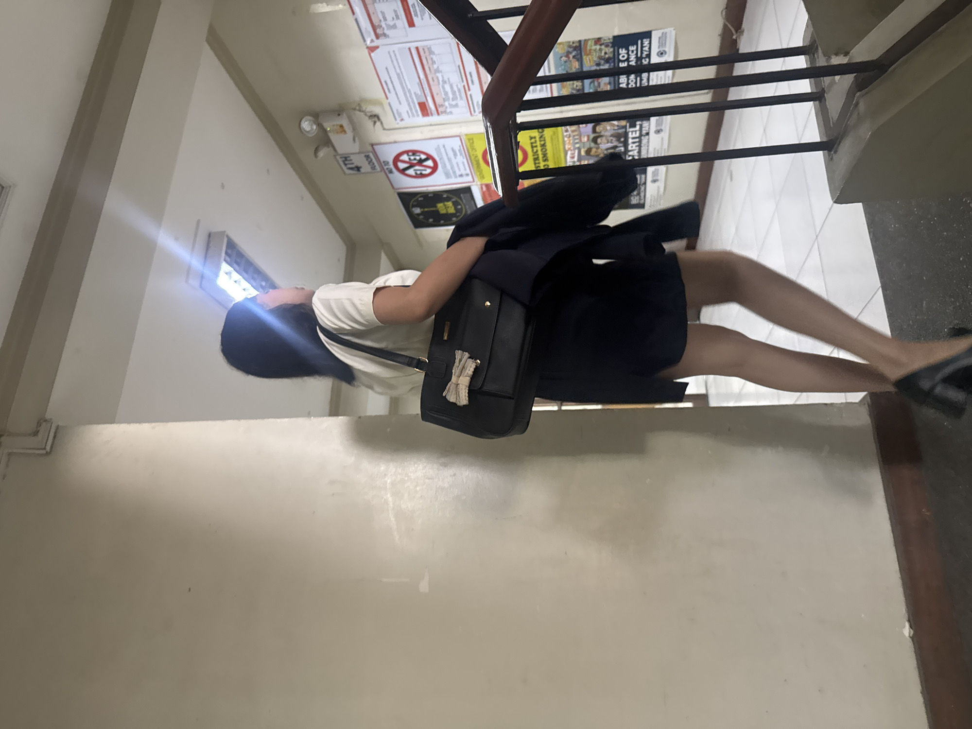 A woman in a white short-sleeved shirt and dark skirt is walking up a staircase. She is carrying a black handbag over her left shoulder and a dark jacket in her right hand. The staircase has a wooden handrail and black metal balusters. The walls are painted a light beige, and there are posters on the wall to the right of the staircase. One poster indicates "4TH FLOOR". The flooring of the staircase is a light-colored tile, and the landing at the top of the stairs is also tiled. The scene appears to be an interior staircase in a building, possibly an office building or a school. The overall lighting is somewhat dim, with a fluorescent light fixture visible on the ceiling. There is no discernible outdoor environment, and the location in Davao City is not indicated by any visual cues in the image.