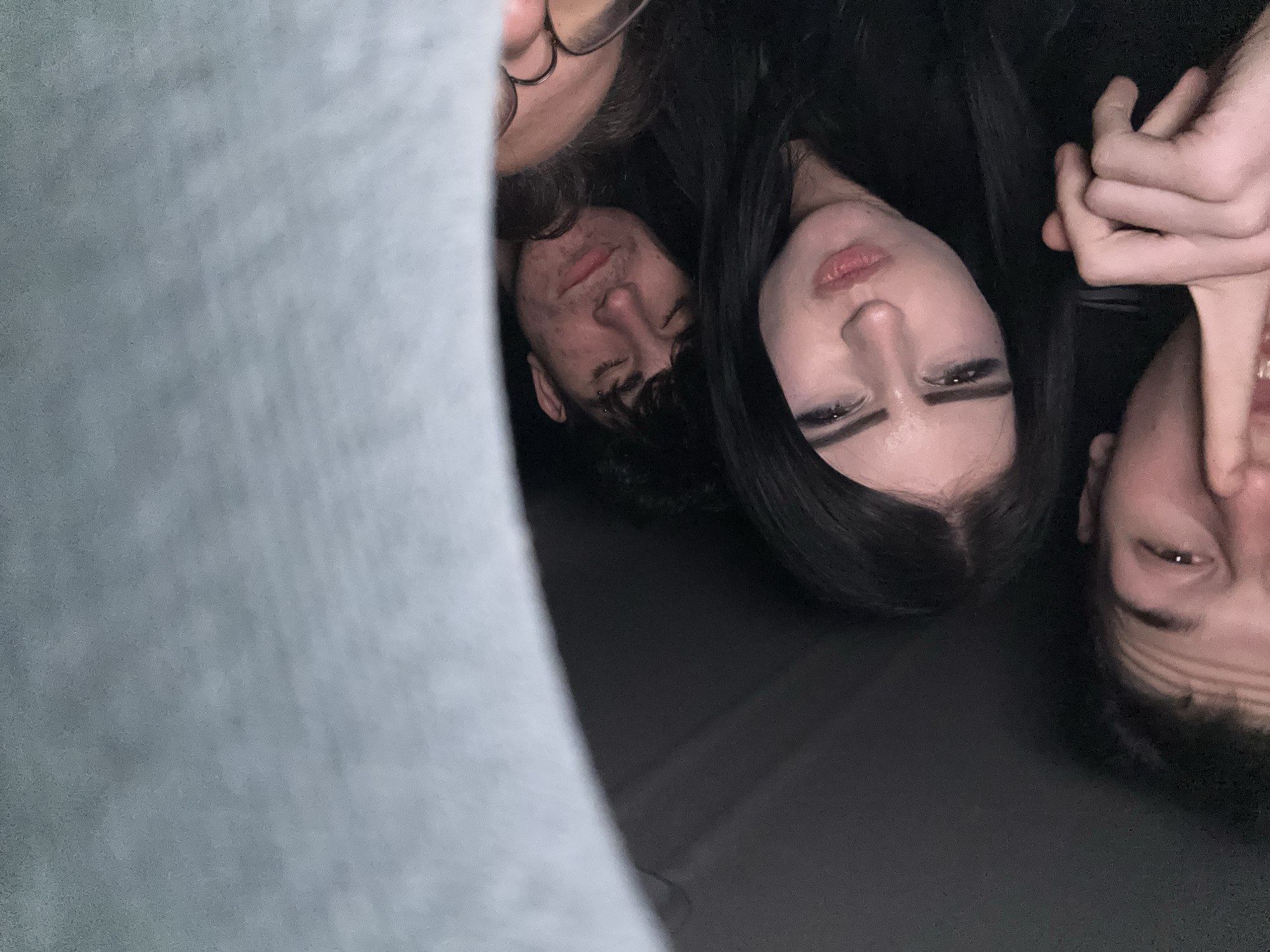 A close-up, upside-down photograph shows three people. A young woman with dark hair and heavy eyeliner is positioned centrally. To her left are two other people, a man with glasses and a man with short dark hair. To the right of the woman is another man with dark hair, his hand is visible in the upper right corner. The photo appears to have been taken indoors. The bottom left corner of the frame features a blurry, light-colored material. The location in Pleven, Bulgaria cannot be identified from this image.