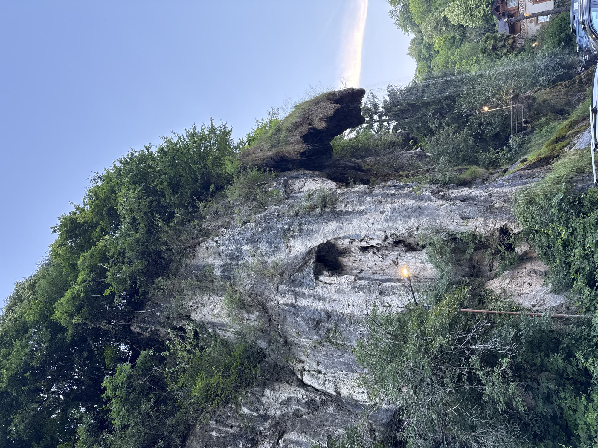 The image depicts a scenic natural landscape in Induno Olona, Italy, featuring a dramatic rock face with a waterfall cascading down its side. The rock formation is covered in lush green vegetation, with trees and moss growing in abundance. The waterfall itself is a prominent feature, with water flowing over the edge and creating a misty spray. In the foreground, a street light illuminates the area, and a few cars are parked below, suggesting this is a public access point or viewpoint. The sky above is a clear blue, with a faint wispy cloud. The overall impression is one of natural beauty and tranquility, with the waterfall adding a dynamic element to the scene.