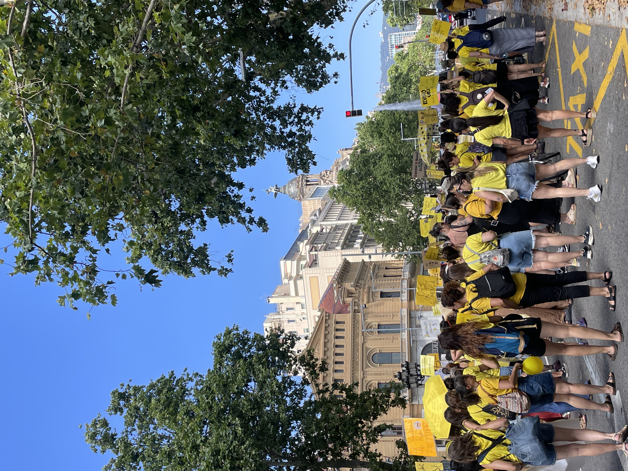 The image depicts a street-level view of a demonstration or protest in Barcelona, Spain. A large group of people, predominantly women, are gathered in the street. Many are wearing yellow tops and carrying yellow signs with text in an unspecified language, suggesting a coordinated event. The background features architectural elements characteristic of Barcelona, including ornate buildings with classical detailing and a dome structure topped with a statue. Trees line the street, and a traffic light is visible in the distance. The ground is paved and marked with yellow lines, including the word "TAXI" painted on the road. The sky is clear and blue, indicating a daytime setting. The people appear to be walking or standing, engaged in a public gathering.