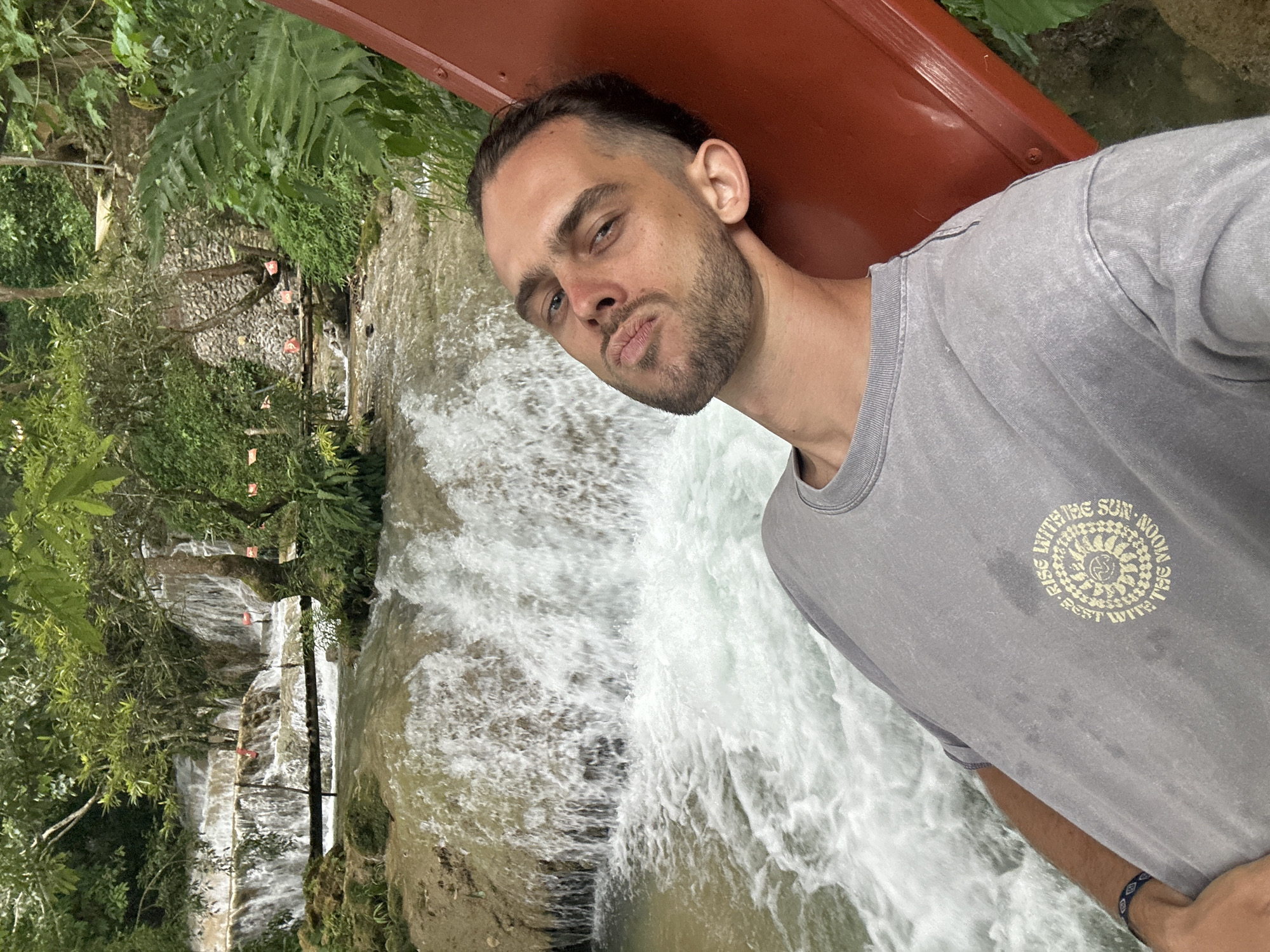 A young man with short, dark hair and a beard is captured in a selfie. He is leaning back on a red slide, with his head tilted back and his face angled towards the camera. He is wearing a grey t-shirt with a circular graphic. Behind him, a multi-tiered waterfall cascades down rocks, surrounded by lush green foliage and trees. The water is turbulent, with white foam indicating a strong current. The overall impression is a natural, outdoor setting.