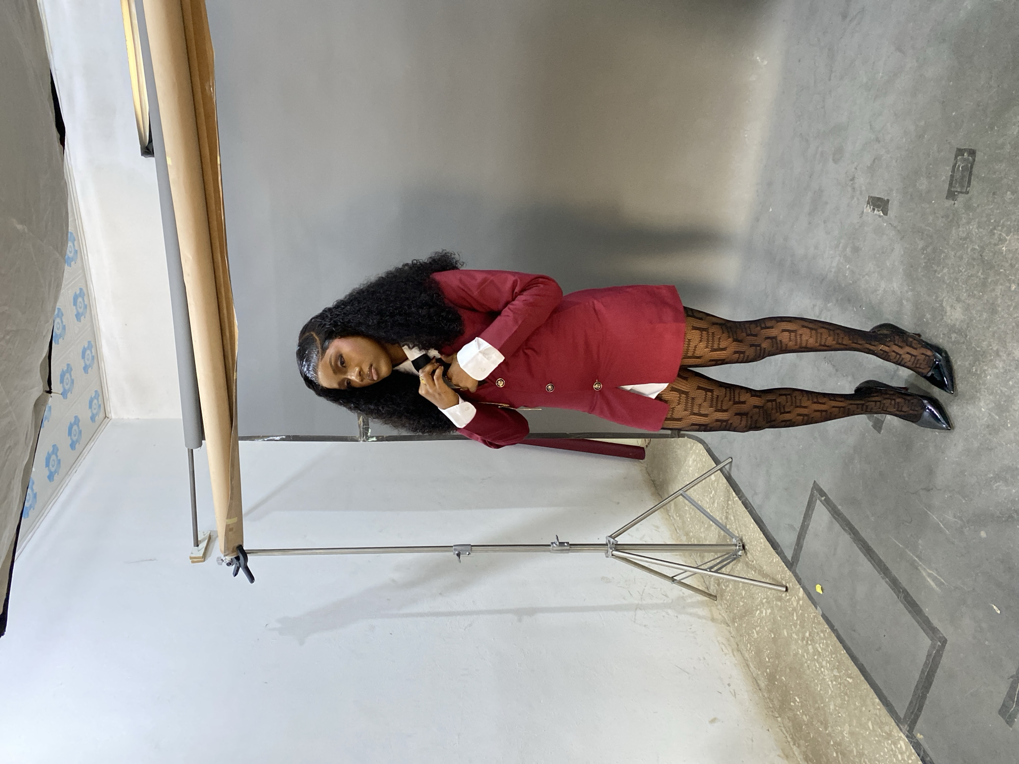 In a studio setting in Gura Top, Nigeria, a single individual, a female with dark skin and dark, curly hair, stands facing partially left while looking leftward out of the frame. She is adjusting a black and white striped tie with both hands at her neck. She wears a dark red blazer-style dress or jacket featuring four gold-colored buttons, a white collared shirt underneath, sheer black tights with a repetitive geometric pattern, and black patent leather pointed-toe high heels.
The background consists of a smooth grey seamless backdrop on the right and a white or light grey wall on the left. Overhead, a white ceiling with light blue patterns is visible, along with a crumpled light-colored fabric. Studio equipment is present, including a silver metal light stand with three legs on the left and multiple rolls of seamless paper (brown/kraft, grey) mounted on a wall bracket above the grey backdrop. The floor is concrete, displaying variations in shade and several rectangular utility access panels. A small, yellow, leaf-like object lies on the floor to the left of the subject.