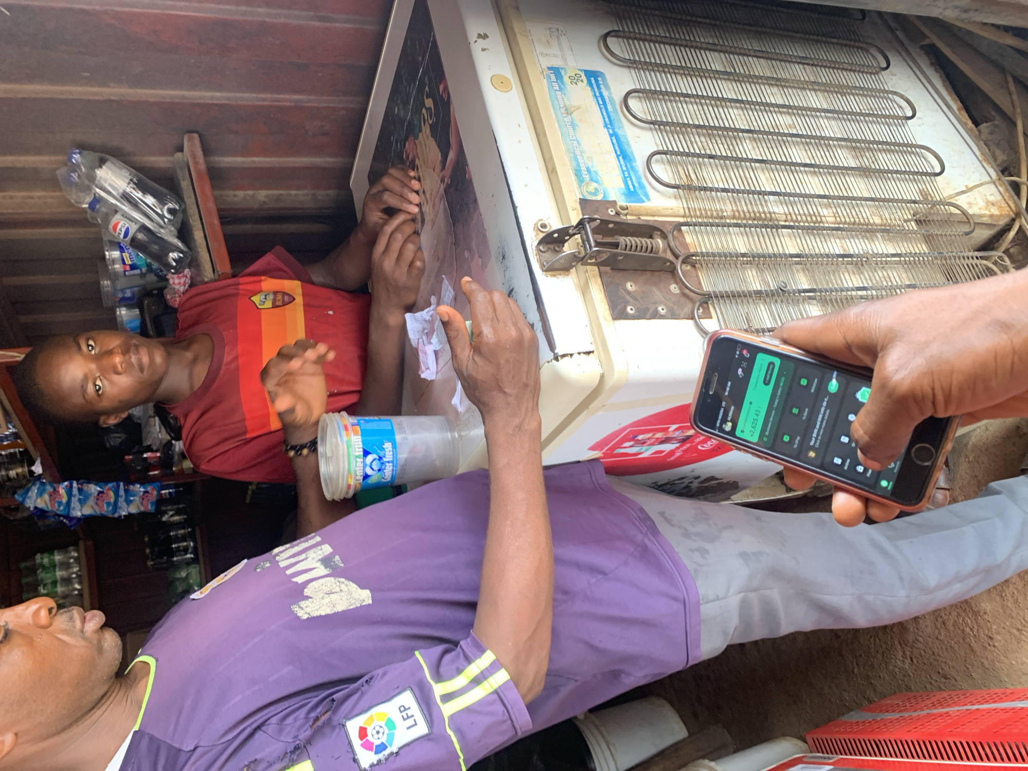 The image shows an interior scene, possibly inside a shop or storage area, in Rafin Par, Nigeria. Two individuals are visible: one wearing a red shirt with a stripe pattern and the other in a purple shirt with a "LFP" patch and yellow striped shoulders. They appear to be interacting with an older refrigerator, which has a sticker that reads "Evangelical Church Winning All Intl." The man in the purple shirt is holding a smartphone, displaying an application with numbers "2,625.43". Various items, including bottles and packaged goods, are present in the background, suggesting a commercial setting. The overall impression is of a casual, everyday scene potentially involving a transaction or interaction related to the refrigerator.