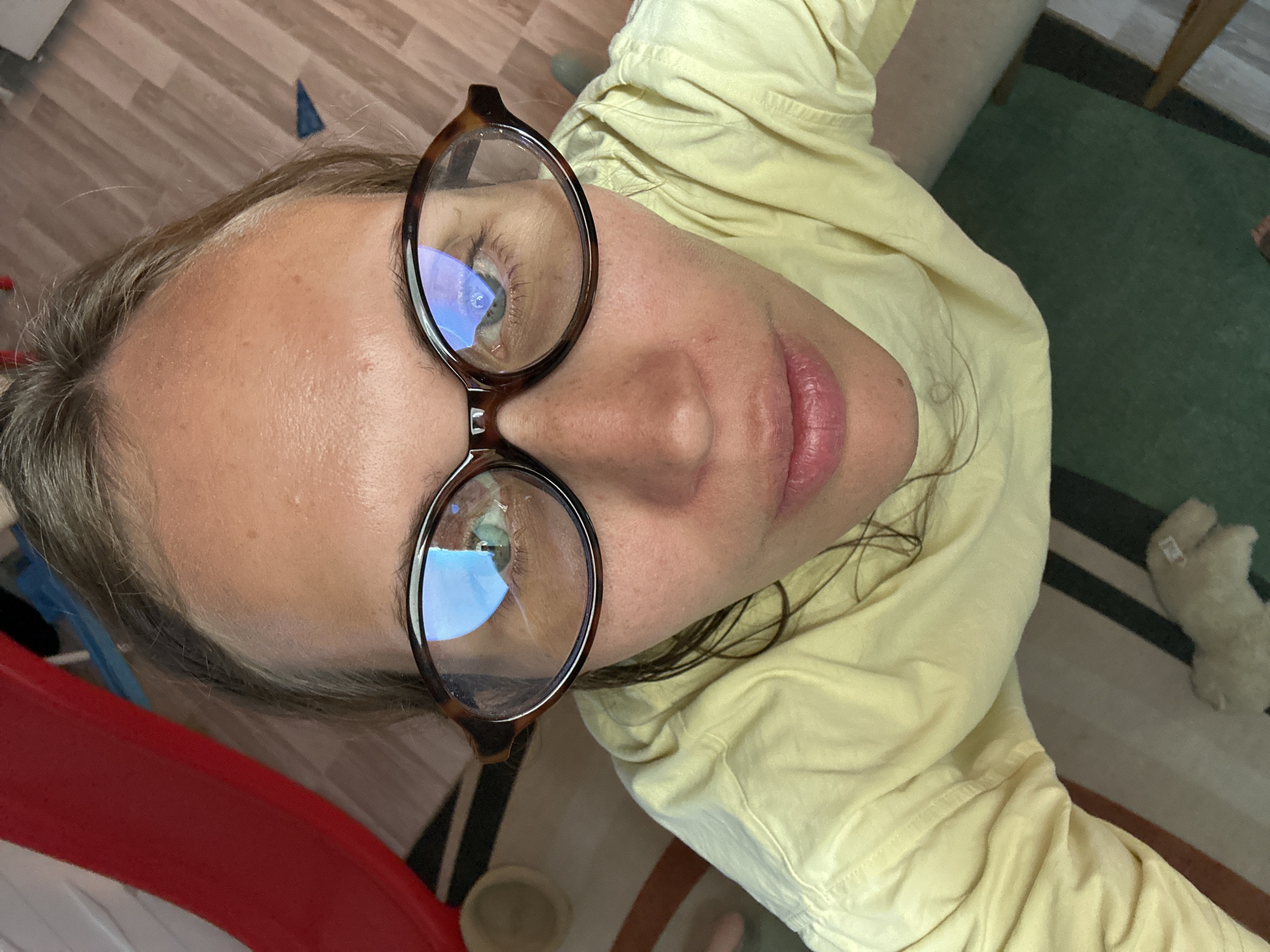 A high-angle close-up shows a person with light skin and light brown hair wearing eyeglasses with brown frames. The person is wearing a yellow garment. The background consists of a wooden floor, a rug with multiple colors, and a stuffed animal. There is no information to conclude this image was taken in Fethiye, Turkey.