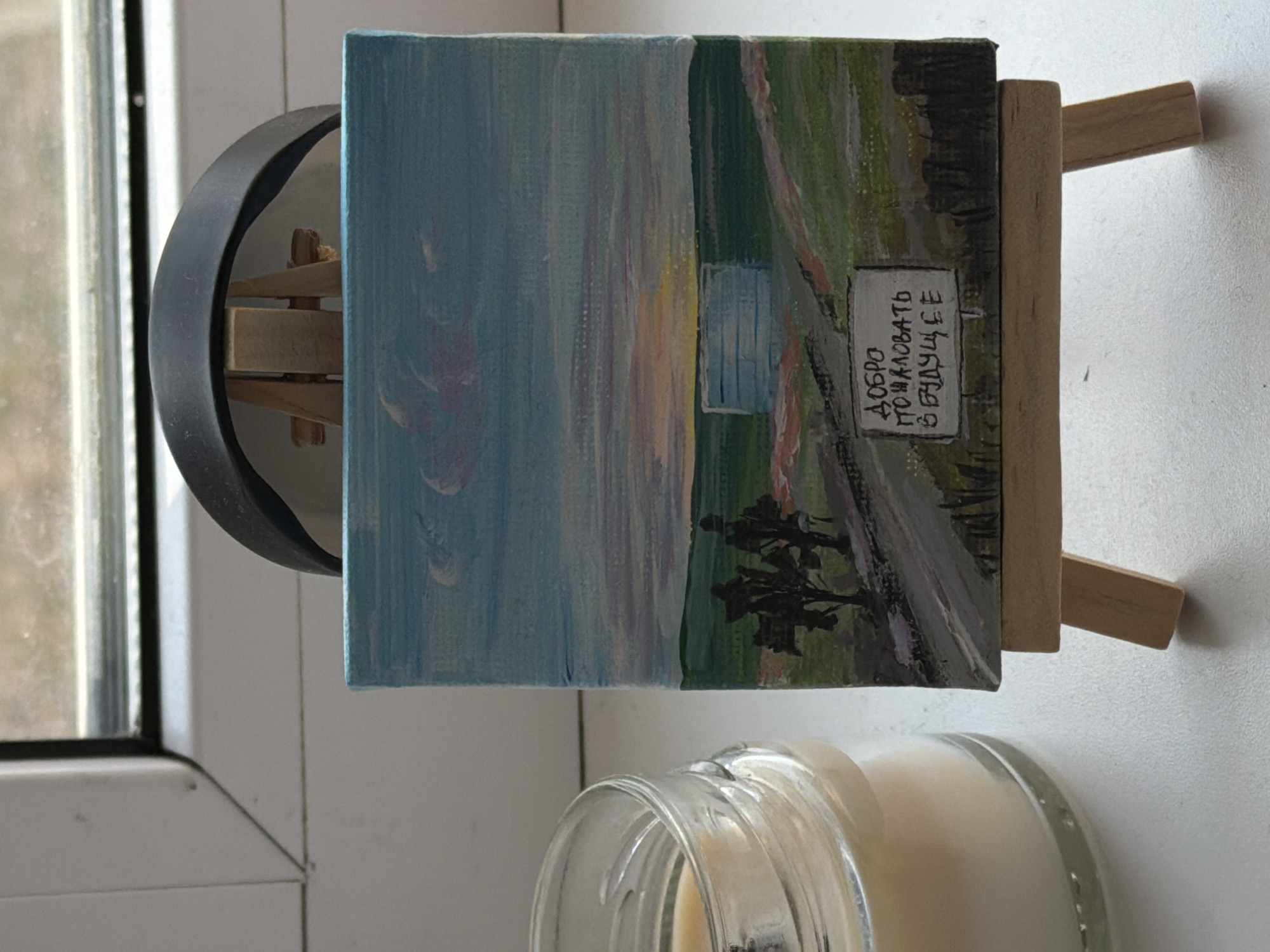 The image depicts a small, landscape painting propped up on a miniature wooden easel. The painting portrays a scene with a cloudy blue sky on the left and a green field on the right. A path with indistinct figures runs along the bottom. A signpost is visible with text in Russian, "Welcome to the future." The painting sits on a windowsill next to a partially visible candle in a clear glass jar. A black ring is also nearby, possibly part of a holder or stand for the easel.
Based on the inscription and the style of the painting, it seems like a local artwork. However, there's nothing in the image to specifically identify the location as the city of Perm, Russia. The general landscape could be found in many regions. More evidence would be needed to confirm its origin.