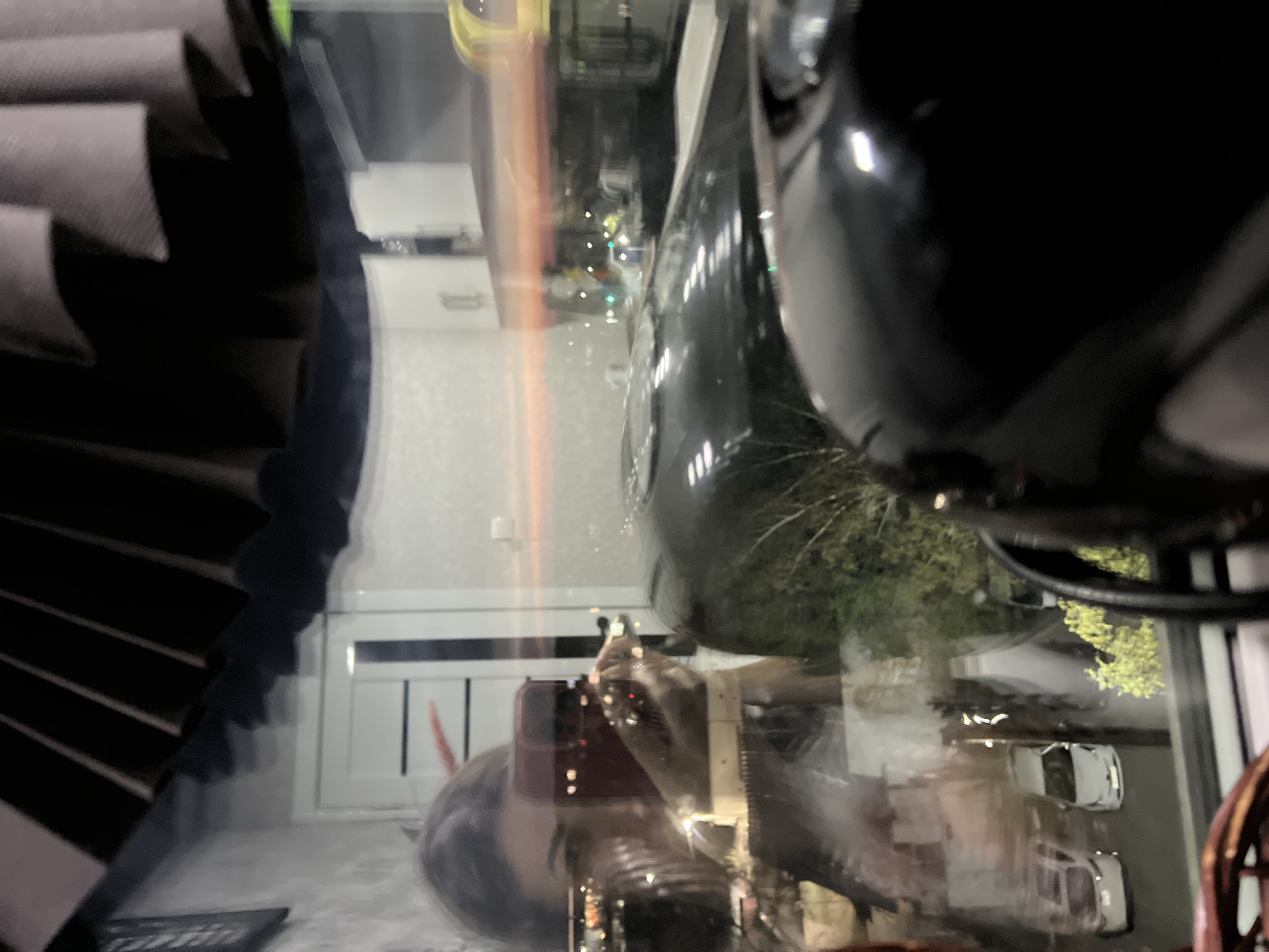 A person is taking a picture through a window. The reflection shows buildings, trees, and cars. The person's phone is visible in the reflection. A black object is prominently in the foreground. The location is an urban area.