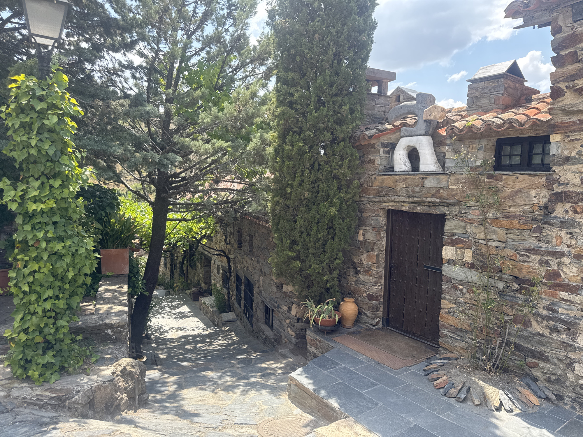 The image depicts a view of a narrow, stone-paved street in Patones De Arriba, Spain. On the right, a rustic stone building with a dark wooden door and a small, black-paned window is prominent. The building features a red-tiled roof and a decorative, abstract stone sculpture above the window. Green foliage and trees are visible throughout the scene, with ivy climbing a lamppost on the left and a tall cypress tree standing near the building. The street is partially shaded by the trees, creating a dappled light effect on the stone paving. In the distance, more stone buildings and vegetation suggest a traditional village setting. The sky is partly cloudy.