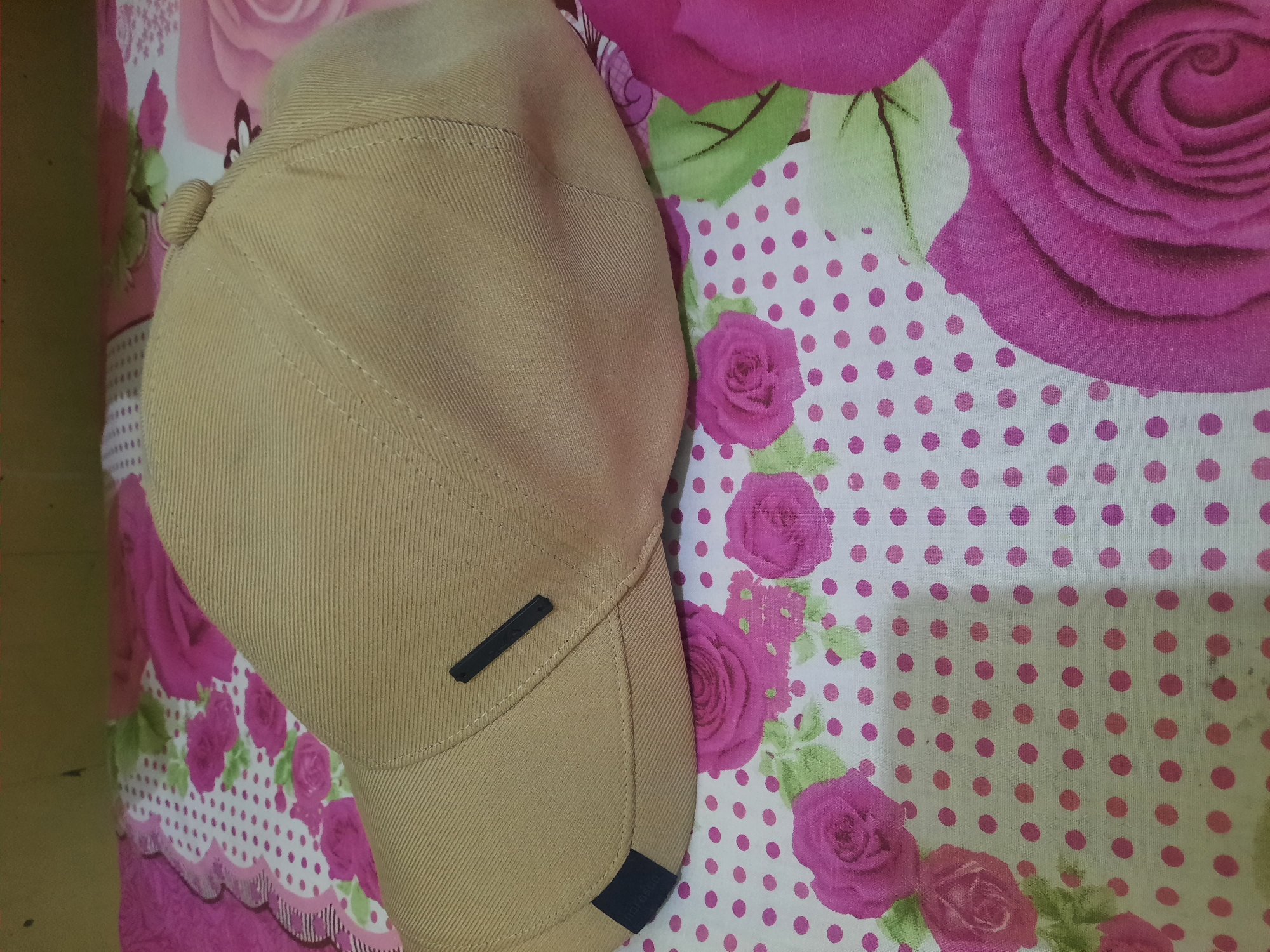A close-up, eye-level shot shows a tan baseball cap on a pink and white polka dot fabric with large pink rose patterns. The cap is positioned diagonally, with its brim facing left, and the crown extending towards the right and top of the frame. A small black rectangular tag is affixed to the side of the crown, and another black tag with some text is on the brim near the left edge. The fabric beneath the cap is a light-colored material with a repeating pattern of pink dots and large, detailed pink roses with green leaves. The background is slightly blurred, suggesting the focus is on the cap and the fabric. The lighting is even, indicating an indoor setting. There are no people or animals visible in the image. The scene appears to be a still life arrangement. The location of Lhokseumawe, Indonesia, is not visually evident from the provided image.