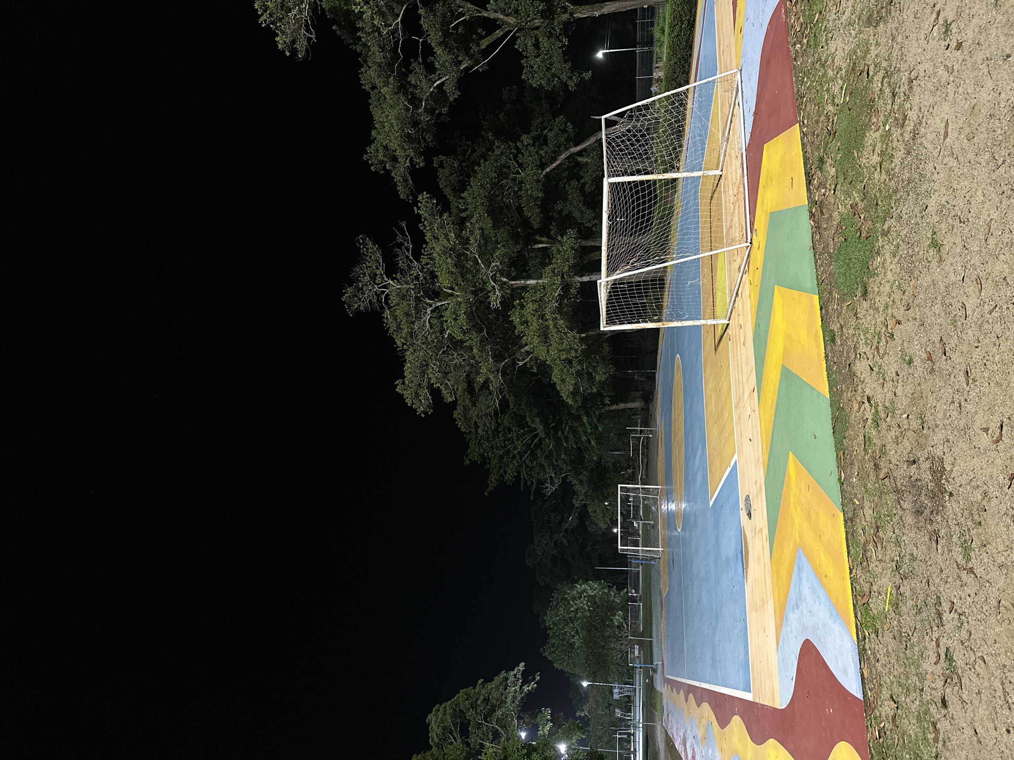 At night, a brightly colored, multi-sport court is illuminated by artificial lighting. The court features distinct sections painted in blue, yellow, and teal, with a red wavy border along the foreground. A soccer goal with a white net is positioned on the court. In the background, trees surround the court, and more sports facilities, possibly basketball hoops, are visible. The scene is set in a park-like area. The location is the public park at the Prince of Songkla University, Phuket Campus, within Phuket City Municipality, Thailand.