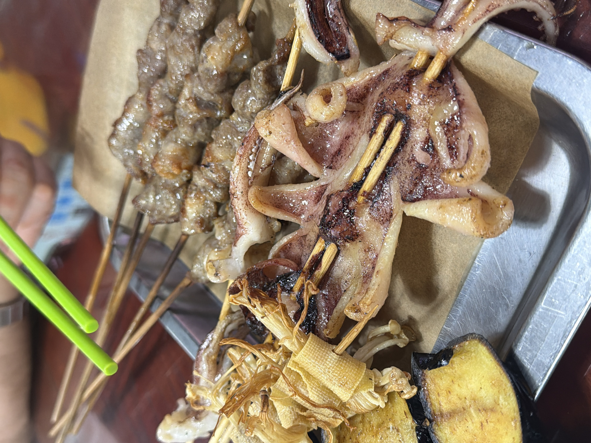 A top-down shot shows a metal tray holding grilled food skewers. On the left, several skewers are stacked with small, glistening pieces of what appear to be meat or organ meat. Adjacent to these are skewers threading a flattened, grilled squid or cuttlefish, marked with dark stripes. Below the squid, more skewers hold what appear to be fried tofu skins, mushrooms, and charred eggplant slices. A pair of green plastic chopsticks rests to the side. The scene likely depicts street food in Dongcheng District, China, implied by the food types and preparation style.