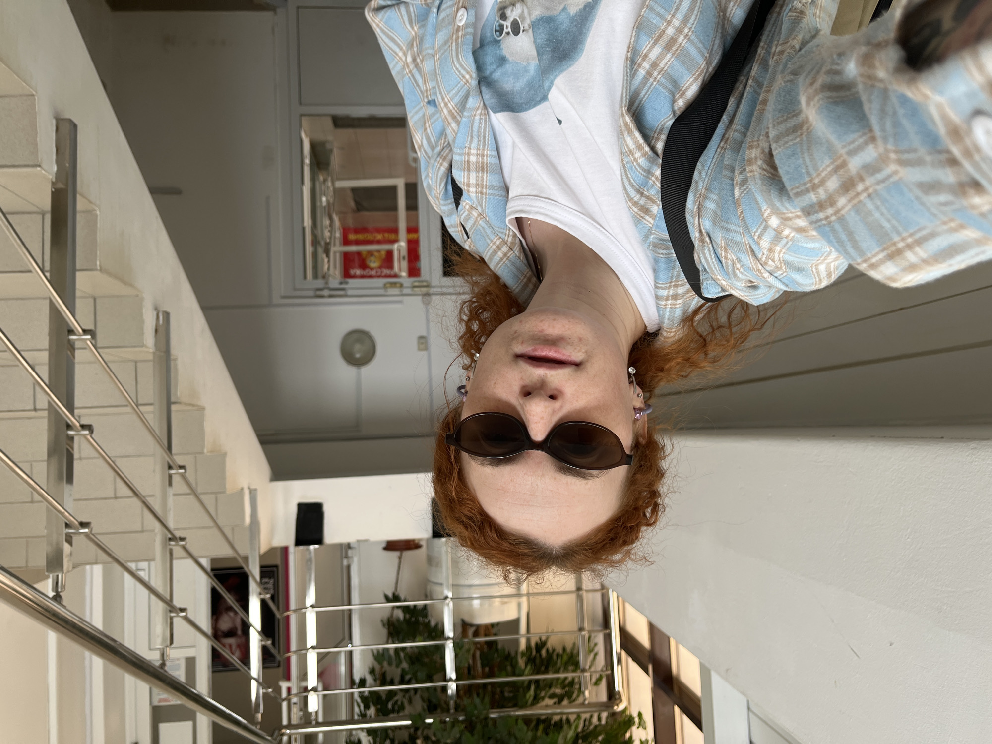 A woman with auburn curly hair and wearing sunglasses, a white t-shirt, and a light blue plaid shirt is the central subject of this selfie. A black strap of some sort of bag is visible over her shoulder. The background includes a staircase with a metal railing, suggesting she might be in a public building or commercial space. Visible through a window behind her is greenery, possibly indicating an indoor plant arrangement or an outdoor area. The scene is located in Chaikovsky, Russia. Additional details are discernible in the background, including a wall, door, and other architectural elements.