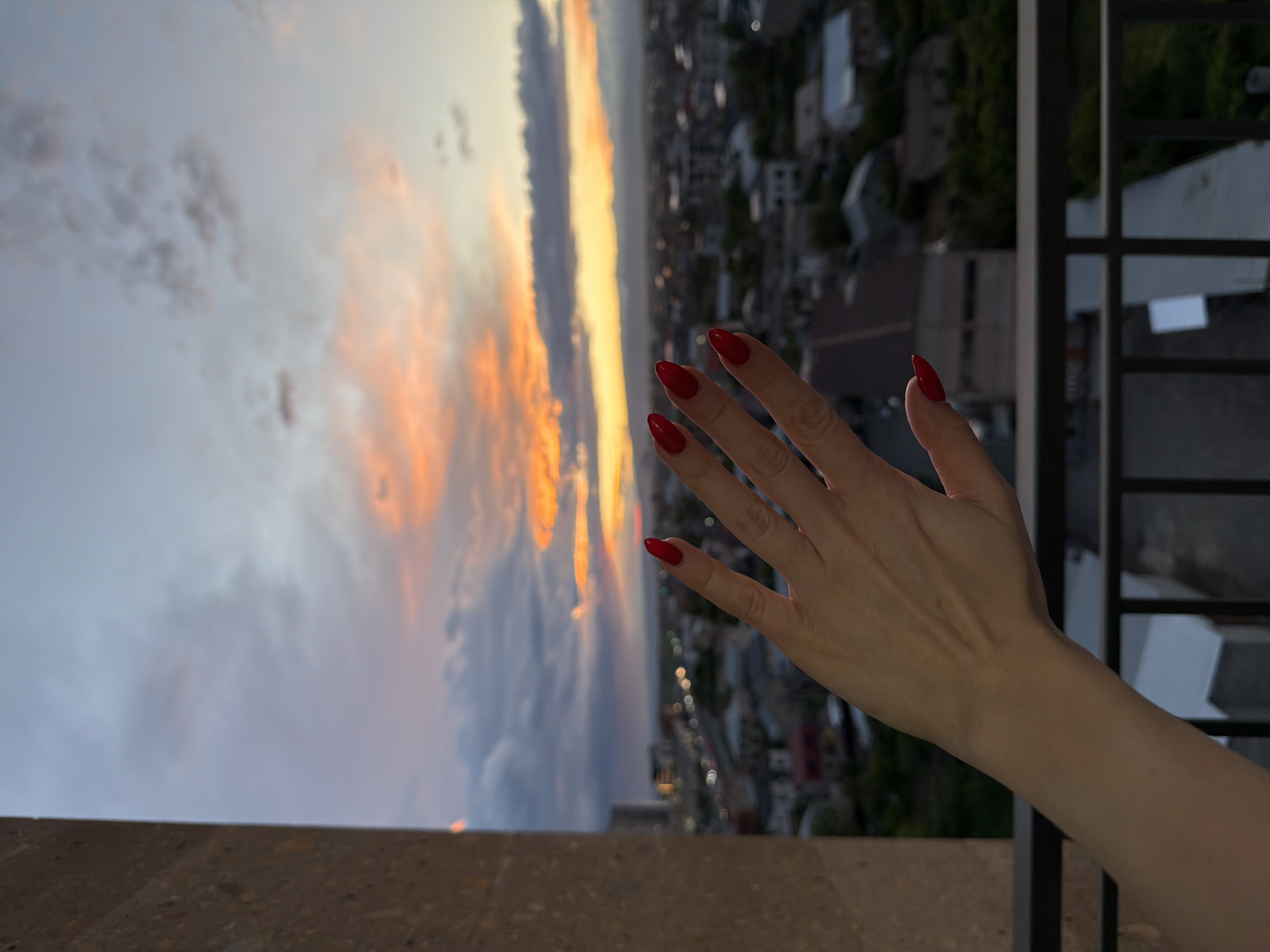 The photograph depicts a partial view of Yerevan, Armenia, captured from an elevated vantage point, likely a rooftop or high balcony. The scene includes a hand with red nails reaching out towards a sunset-lit sky, suggesting a personal moment of observation. The city's buildings are visible in the background, blurred to emphasize the sunset and hand. A section of a balcony railing is visible to the right. The primary focus is on the atmospheric conditions and the individual's interaction with the view. The image suggests a quiet, contemplative moment.