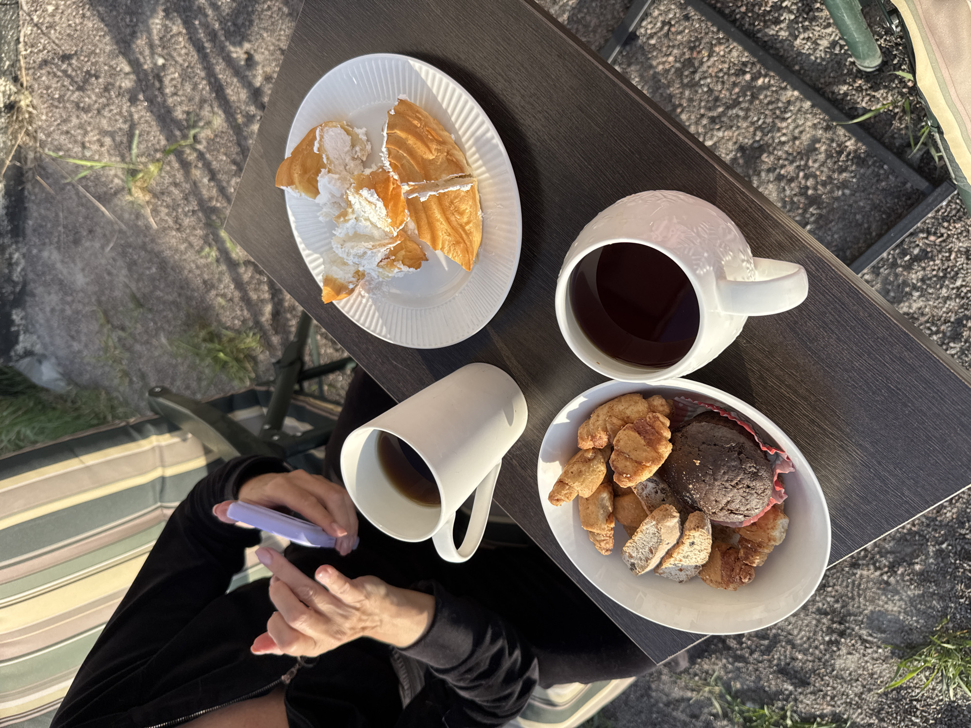 The image presents an outdoor setting, possibly in Steklyannyi, Russia, though this is speculative. A person is seated on a striped chair, clad in a black track suit, and holding a lavender-colored electronic device. Positioned on a dark, wood-look side table are two cups of dark liquid, likely coffee or tea. One cup is tall and plain white; the other is a larger, white mug with an embossed floral design. Two white plates hold food items. One plate contains a sliced pastry filled with cream, while the other contains an assortment of pastries and a large muffin. The ground is gravelly with sparse patches of grass.