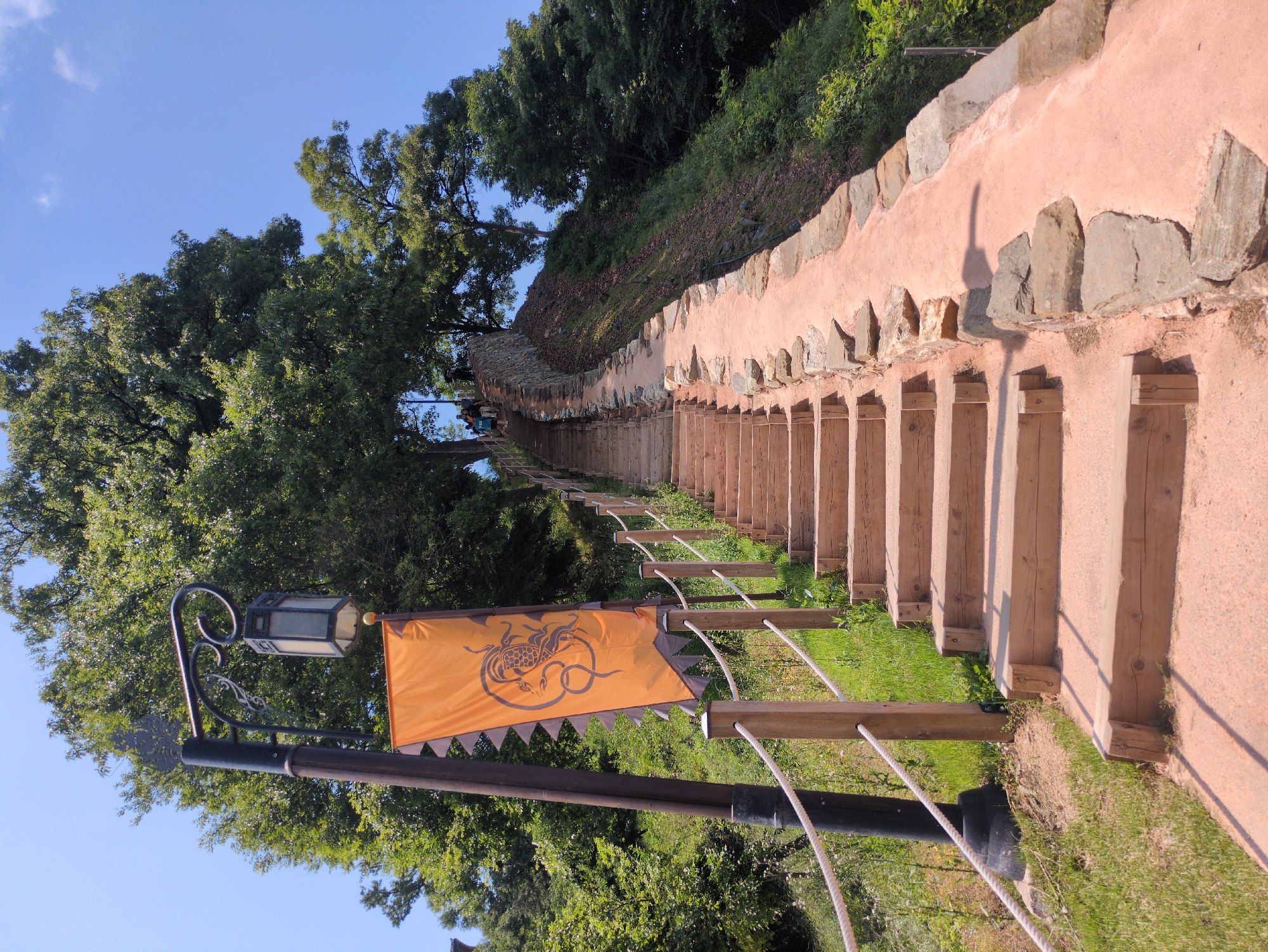 The image depicts a long staircase leading uphill, in Gongju-si, South Korea. The steps appear to be made of light-colored wood, with a pale pink or light brown pathway on either side. A wall runs along the right side of the staircase, constructed with pinkish material and inlaid with stones. A rope railing, supported by wooden posts connected to a black metal structure, runs along the left side. A lantern with an attached orange flag bearing a serpentine design is visible on the left, near the beginning of the staircase. Trees and greenery line both sides of the path, with a clear blue sky visible above. The staircase stretches into the distance, suggesting a significant incline. People are partially visible in the distance at the top of the stairs.