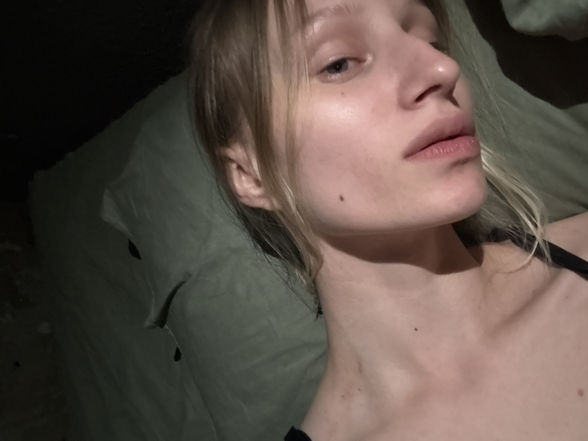 A fair-skinned individual with blonde hair is lying down on a green surface, possibly a pillow or cushion. The setting appears to be indoors, with a dark or dimly lit background. The individual has a mole on their cheek and another on their neck. The person is wearing what appears to be a black strap, possibly from clothing, visible on their shoulder. The location is unknown and there is no mention of Volkovka, Russia.