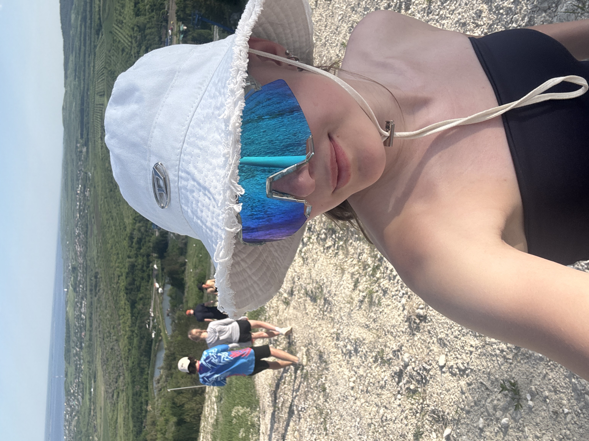 A selfie-style photograph shows a person wearing a white bucket hat and sunglasses with blue lenses. The person is also wearing a black top. In the background, there is a gravel surface with green vegetation. A group of three people is visible further away, walking on a path. The background shows a landscape with trees and a body of water, suggesting an outdoor environment. The city of Khvalynsk, Russia, is not explicitly identifiable in the image.