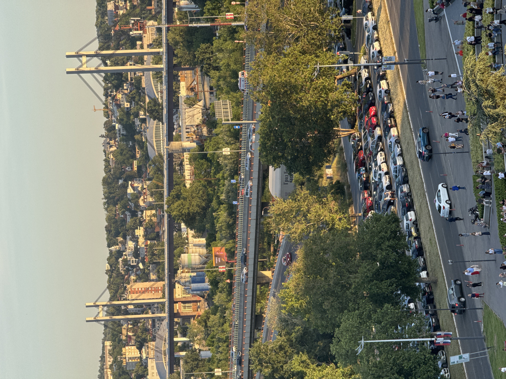 Here is a description of the image:
The image shows an elevated view of a scene in an urban area, likely a city. In the background to the left is a bridge with several suspension towers. A highway runs in the middle of the frame, with visible traffic. Parallel to the highway on the right side of the image is another road or street, which is lined with parked cars. Additionally, a pedestrian path with people is also present next to the cars. Trees and greenery are interspersed throughout the scene, especially in the area between the highway and the secondary road/pedestrian path. The image suggests a mixed-use area combining transportation infrastructure, residential or commercial areas, and green spaces.