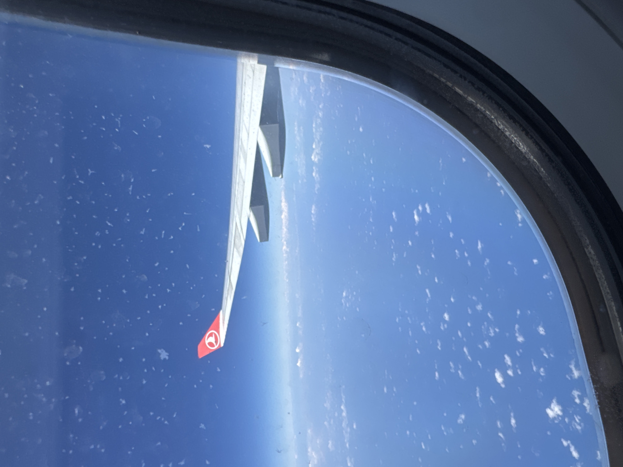 The image shows an airplane wing, viewed from inside the aircraft through a window. The wing has a red tip with a white emblem on it. The window frame is visible. Outside the window, there is a blue sky with some cloud cover. The picture appears to be taken mid-flight. The location, Tambon Sa Khu, Thailand, cannot be verified from the image.