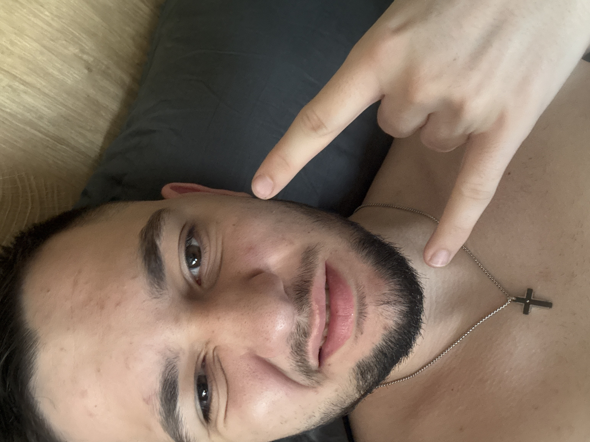 A man with dark hair and a beard is lying on his back, possibly on a gray pillow, with light wood flooring visible in the background. He is wearing a silver chain necklace with a cross pendant. He is making a hand gesture resembling devil horns with his right hand, positioning the index finger near his cheek and the little finger near his neck. The setting is not definitively identifiable as Pernik, Bulgaria; no landmarks or contextual clues are present in the image to confirm this location.