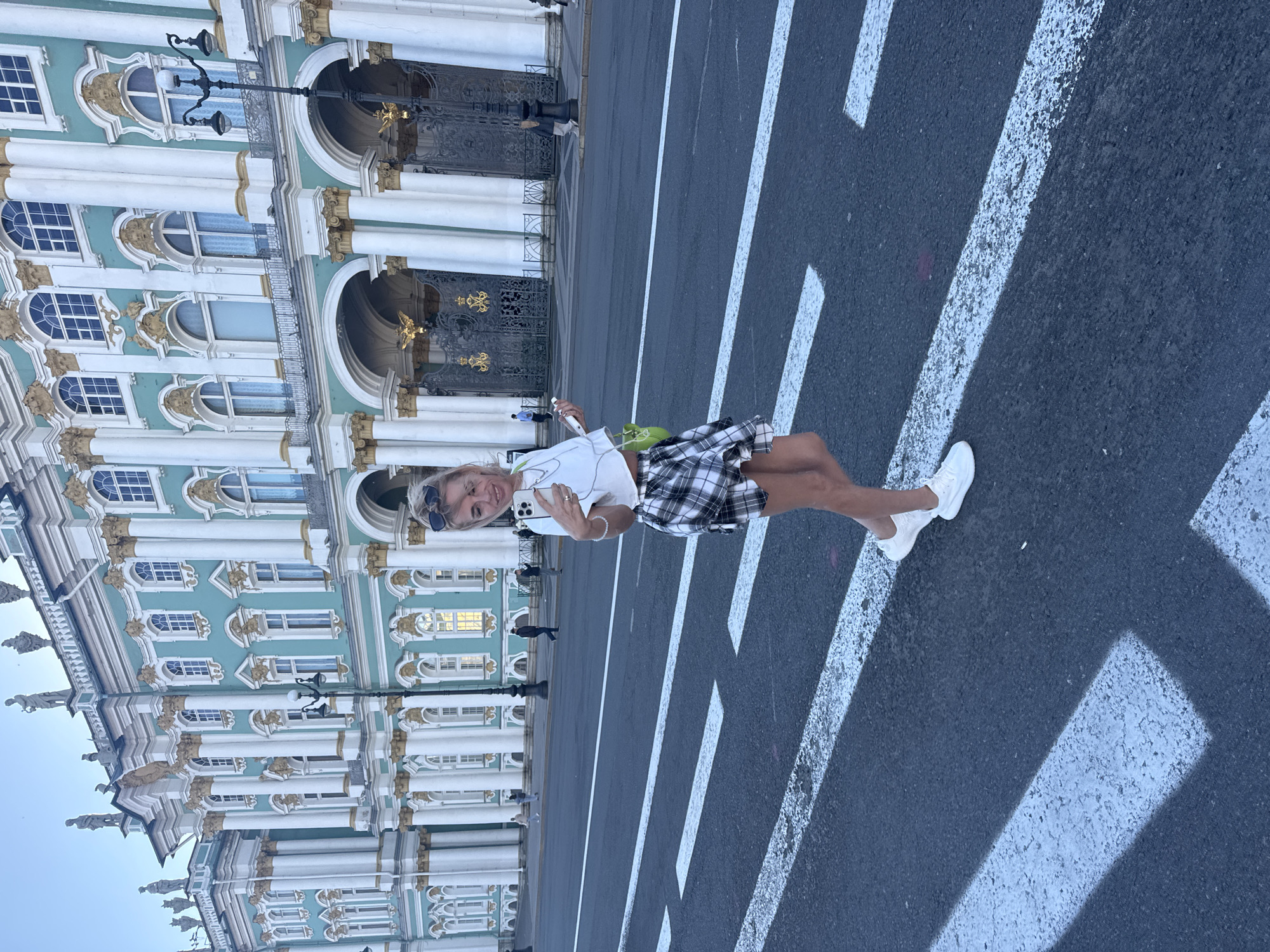 A young woman with blonde hair and sunglasses is taking a selfie while standing on a crosswalk in front of the Winter Palace in St. Petersburg, Russia. She is wearing a white crop top, a black and white plaid skirt, and white sneakers. A lime green bag is slung over her shoulder. The Winter Palace, with its distinctive turquoise facade and white columns, dominates the background. Several people are visible walking in the distance. The sky is clear and overcast.