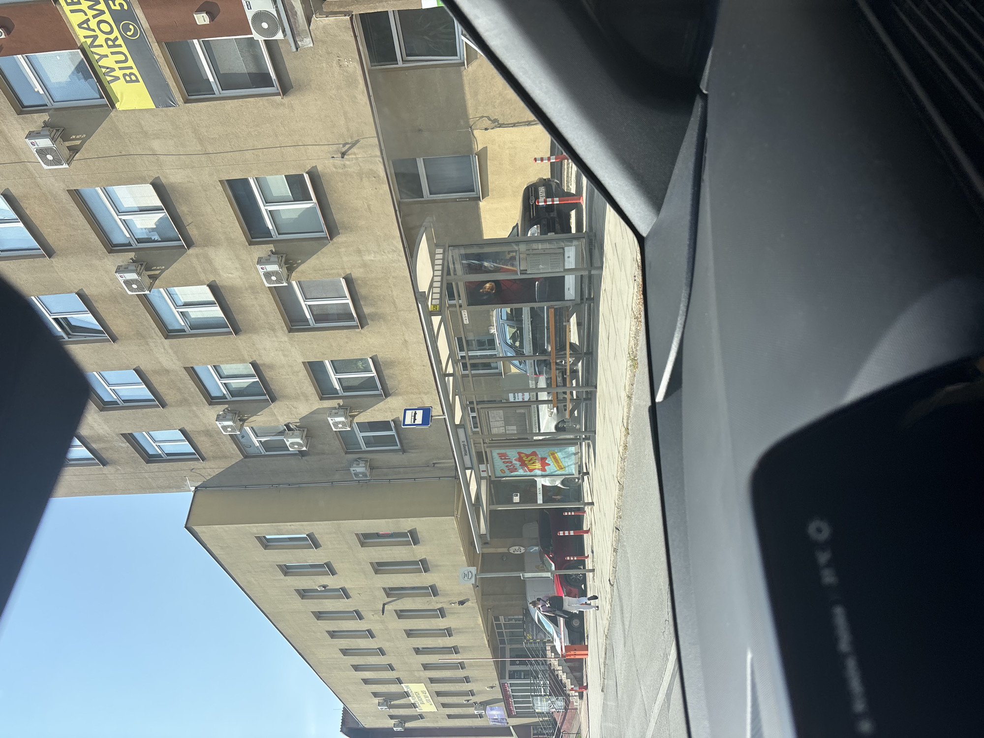 The image depicts a street scene in Krakow, Poland, likely on a sunny day. In the foreground, the interior of a vehicle is visible, with a dashboard displaying a temperature of 23°C. The main focus is a bus stop situated in front of a multi-story building. The building appears to be an older, light brown structure with numerous windows and external air conditioning units. Some windows have reflections, obscuring the interior. A yellow banner with Polish text, likely an advertisement for office rentals, is attached to the building.
The bus stop is a covered structure with a glass enclosure, displaying advertisements, one of which mentions "103.8 FM KISS". A bus stop sign is visible. Outside the bus stop, several vehicles are parked or waiting. These include a red car, a black SUV, and a white van. Orange and white traffic cones are placed along the sidewalk.
A person is seen walking away from the camera on the sidewalk, wearing light-colored pants and a backpack. The street appears to be paved, and a sidewalk runs alongside it. The sky is clear and blue. The overall impression is of an urban environment during daylight hours.