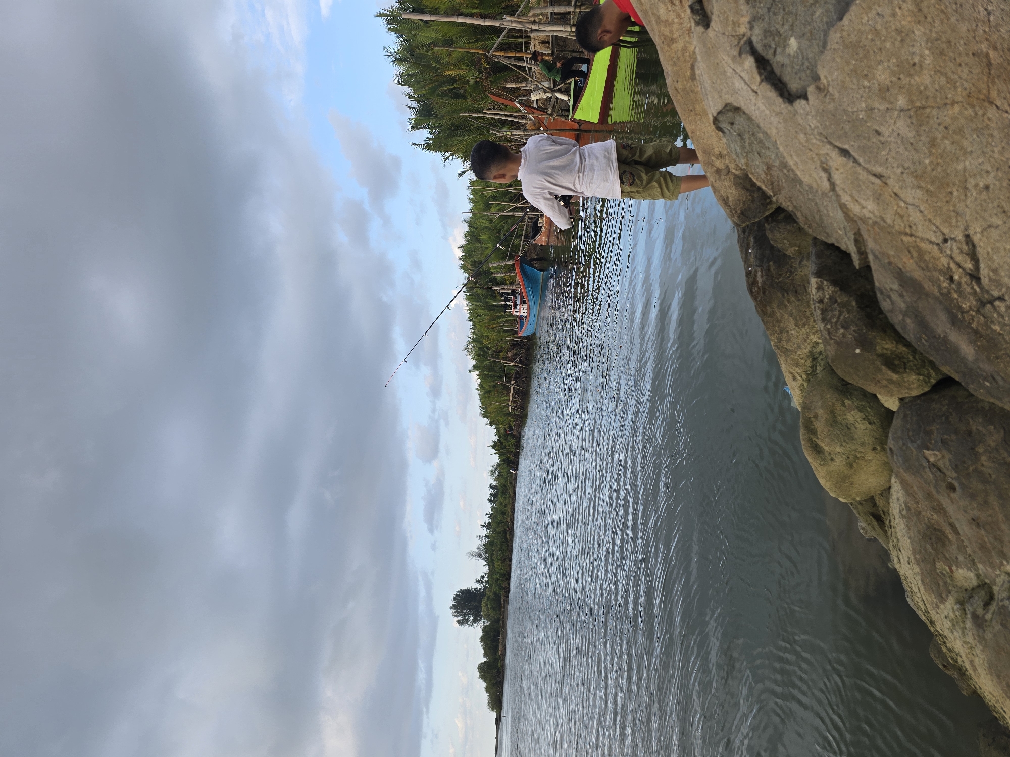 In Reudeuep, Indonesia, on a cloudy day, a young boy is seen fishing by a body of water. He stands on a rocky embankment, holding a fishing rod with his back to the camera. In the background, several boats are moored along a shore lined with palm trees. One boat is blue, another appears orange, and a third is bright green. Another person, wearing a red shirt, is partially visible on the right side of the frame. The water is calm with ripples on its surface, and the sky is filled with gray clouds.