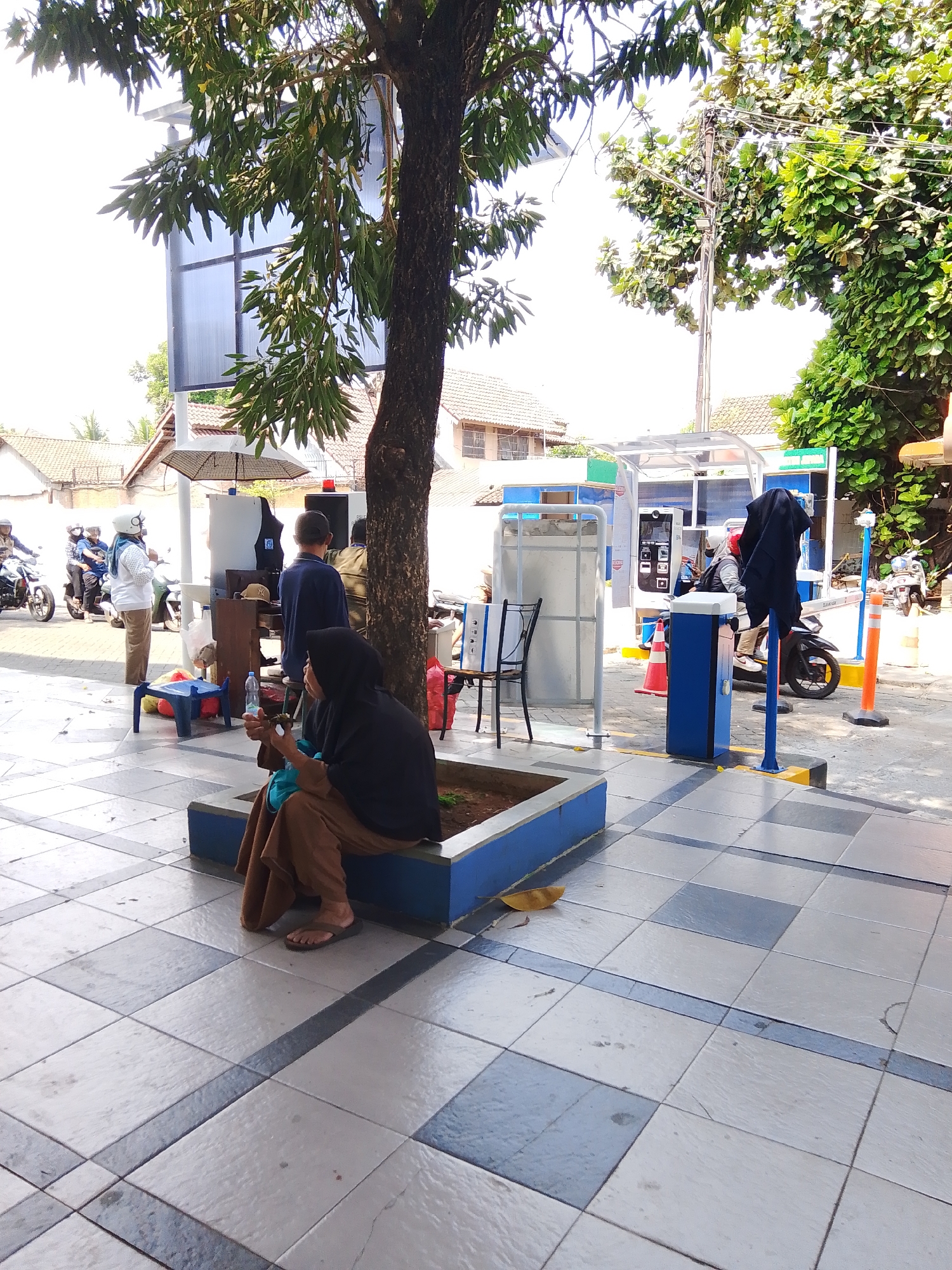 The image depicts an outdoor scene, likely a public area in Potroyudan, Indonesia, possibly near a charging station or amenity area. In the foreground, a person wearing a black hijab and brown attire is seated on a raised planter box, holding a mobile phone. The planter box is lined with blue and has soil inside. The ground is paved with square tiles in a checkered pattern of white, grey, and some darker blue-grey lines. Behind the seated individual, a large tree trunk dominates the mid-ground, with its branches and leaves partially obscuring the sky and background structures.

To the left of the tree, several people are visible. One person in a white shirt and beige pants stands near a white structure. Several individuals on motorcycles are also present; one in a white helmet and blue shirt is on a motorcycle, and another person in a black helmet is also on a motorcycle. Further back, a white umbrella is set up, and another person in a dark blue shirt is standing.

In the background, a modern-looking structure with a translucent roof and white supports appears to be a covered waiting area or service point. It has a digital display or panel on its side. Orange traffic cones and blue bollards are strategically placed around the area, suggesting a controlled or designated space. Beyond this structure, residential buildings with tiled roofs are visible, indicating the setting is within a populated urban or semi-urban environment. Lush green trees are present on the right side of the image, adding to the natural elements of the scene. The lighting suggests it is daytime, with bright sunlight casting shadows.