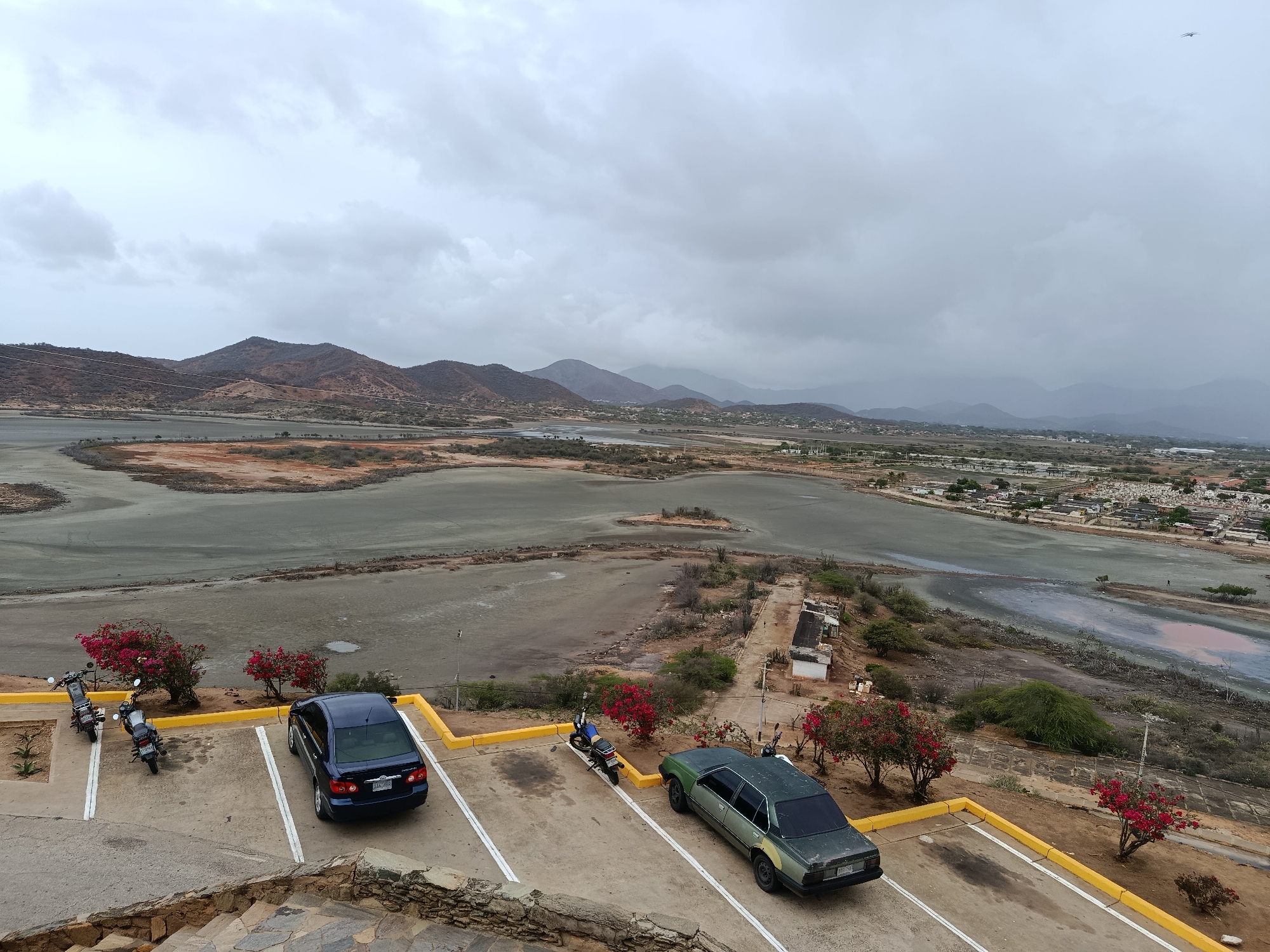 This media file is an eye-level, high-angle shot of a parking area overlooking a salt flat and a distant town. The scene is set on an overcast day, with heavy, grey clouds dominating the sky, creating a diffused light that casts no strong shadows. The landscape is characterized by arid, reddish-brown hills and mountains in the background, with a vast, shallow body of water, likely a salt flat, in the midground. The water's surface appears somewhat textured and reflects the muted sky.
In the foreground, a paved parking lot is marked with white lines. Two dark blue sedans are parked, one in a designated spot. A faded, dark green sedan is also parked in a spot, with visible repairs or discoloration on its rear fender. Beside the blue sedan, three motorcycles are parked in a line. Vibrant pink flowering bushes are strategically placed around the parking area, providing a splash of color against the otherwise muted tones of the landscape. A stone staircase is visible on the lower left of the frame, suggesting the viewpoint is elevated.
In the distance, a town with low-rise buildings is nestled near the edge of the salt flat, extending towards the hills. A small road with a few scattered structures runs along the edge of the water. The overall mood is quiet and somewhat somber, due to the overcast weather. No people are clearly visible, but the presence of parked vehicles suggests recent or current human activity in the area. The location is identified as Juan Griego, Venezuela. A license plate on the blue sedan is partially visible, displaying text that is difficult to discern clearly. The green car's license plate is also visible and readable as "FAS-158".