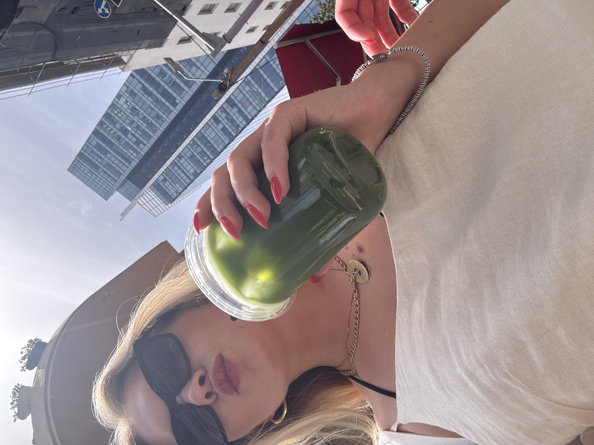 A woman with blonde hair and sunglasses is holding a cup of green juice. She is wearing a white linen dress and gold jewelry. A high-rise building is visible in the background. The location is likely in the city of Tel Aviv, Israel.