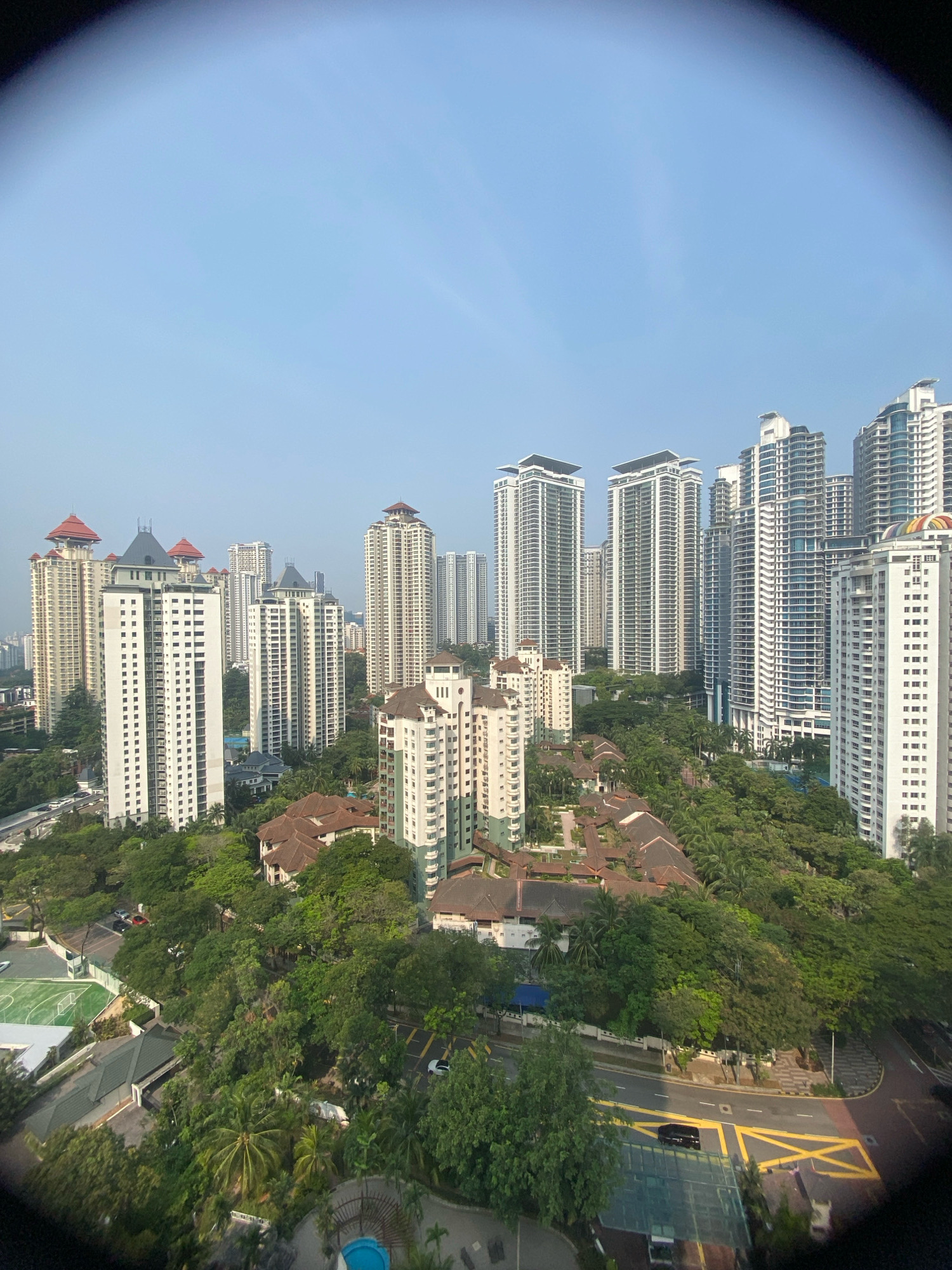 This is a high-angle, wide-view, exterior shot of a dense urban landscape in Kuala Lumpur, Malaysia, likely taken from a tall building. The image is slightly distorted by a fisheye lens effect, particularly around the edges. The scene is dominated by numerous high-rise residential buildings, many of which are modern skyscrapers with sleek, white facades and balconies. These are interspersed with lower-rise buildings featuring red-tiled roofs, characteristic of some residential areas in the city.
Extensive greenery, including mature trees and lush vegetation, fills the spaces between the buildings and surrounds a sports field with white lines visible. Roads with visible lane markings and at least two cars are present, indicating active transportation routes. The sky is a clear, pale blue, suggesting it is daytime, likely morning or late afternoon given the soft lighting. The overall impression is of a well-developed, modern city with a significant amount of green space integrated into its urban fabric. The specific location is likely in an affluent residential district, given the scale and architecture of the buildings and the presence of amenities like the sports field. Without further identifying landmarks, pinpointing the exact neighborhood is not possible.