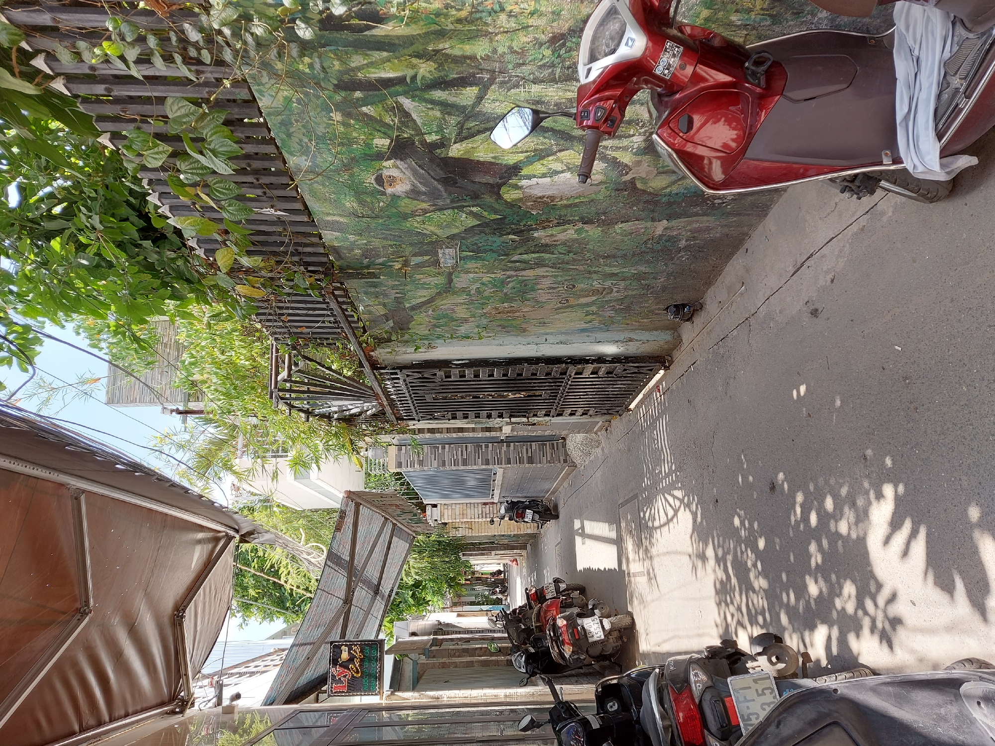 The image shows a narrow alleyway in the city of Đà Nẵng, Vietnam. The alley is lined with buildings on the left and a wall with a mural on the right. Several motorbikes are parked along the left side of the alley. The mural on the right wall depicts a monkey in a forest setting. The ground is paved with asphalt. The sky is visible at the top of the image and appears to be clear. The lighting suggests it is daytime. There are no people visible in the image. The overall scene is that of a typical urban alley in Vietnam.
