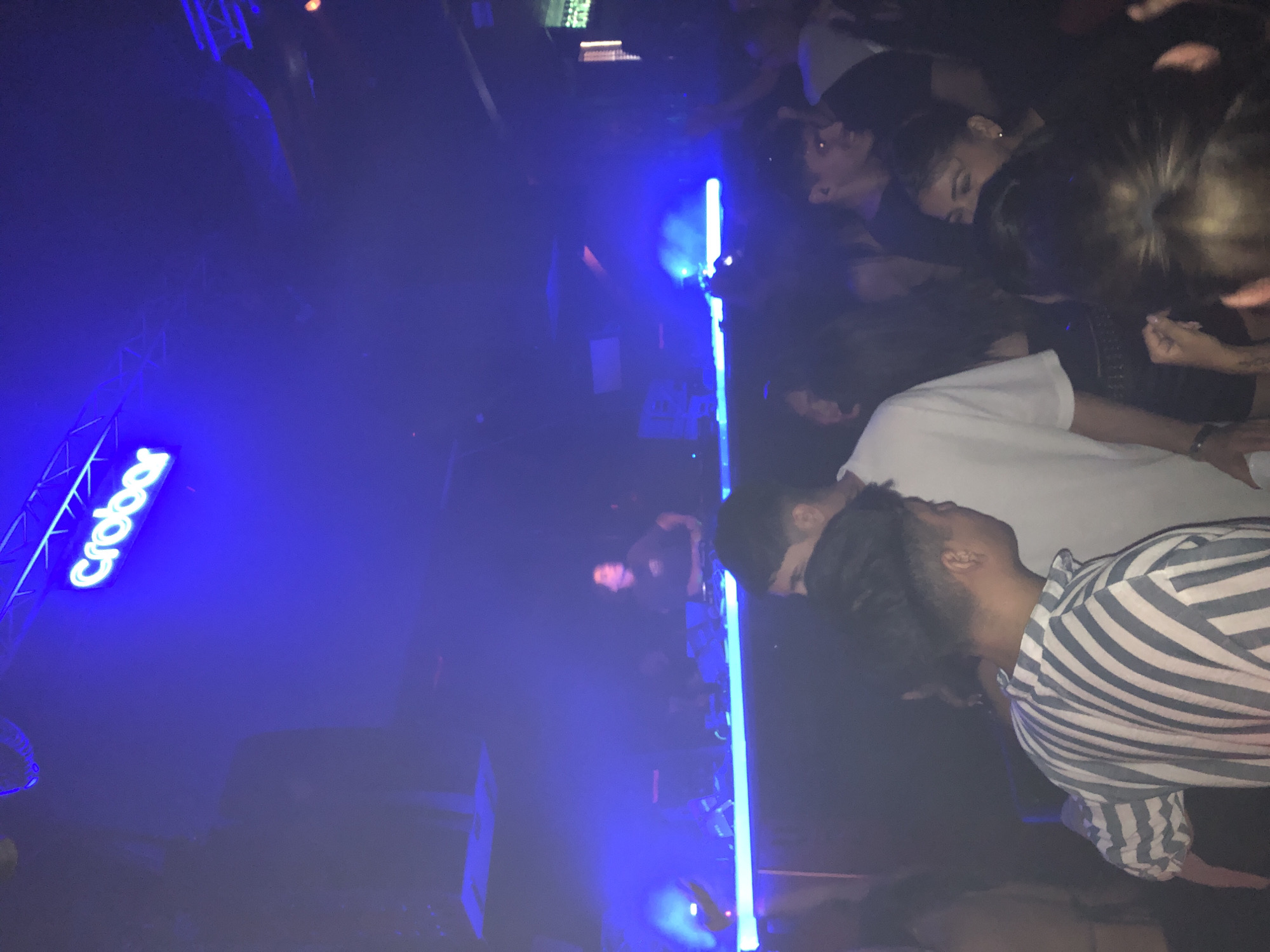 Here's a forensic description of the image:
**Scene:** Indoor environment, likely a nightclub. The lighting is primarily blue, creating a dark and hazy atmosphere.
**Objects:** A blue light bar is visible, emitting a strong glow. Signage with the word "Crobar" is lit up in blue neon.
**People:** Multiple individuals are present, suggesting a crowd. Their faces are not clearly visible due to the lighting and focus. Some are wearing casual clothing like a white t-shirt and a striped shirt.
**Actions/Interactions:** The people seem to be in close proximity to each other.
**Location:** This image was taken at Crobar Nightclub in the city of Buenos Aires, Argentina.