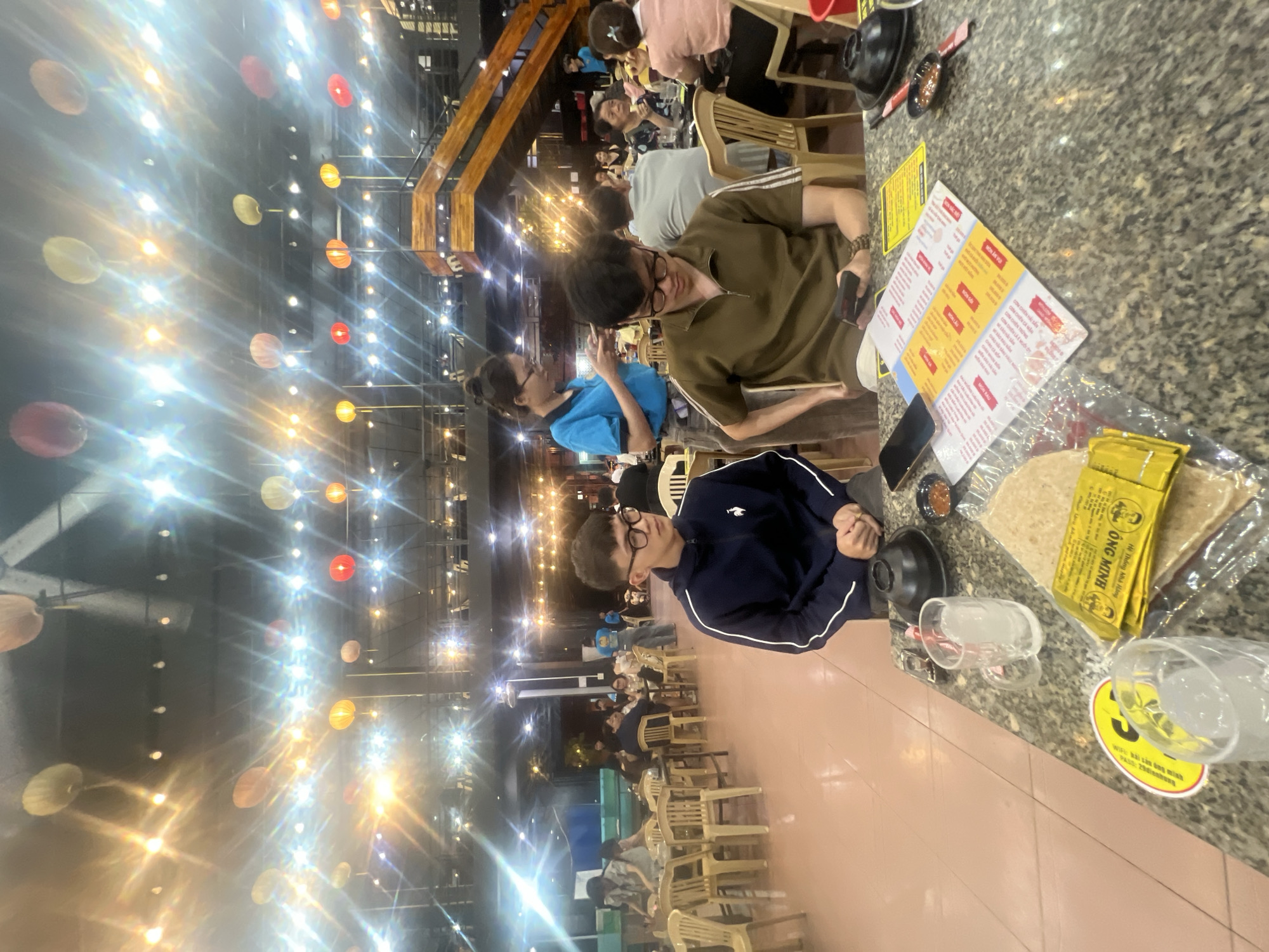 The image captures a lively evening scene inside a restaurant or casual eatery in Quy Nhon, Vietnam. The space is illuminated by numerous small, bright artificial lights strung across the ceiling, interspersed with decorative paper lanterns in red, yellow, orange, and white/beige hues, creating a warm ambiance.
In the foreground, a speckled granite table is seen with two young men and a woman seated around it. One young man, wearing a dark blue jacket with white stripes and glasses, looks towards the left. Beside him, another young man in a brown polo shirt and glasses is looking down, seemingly at his phone or the menu. A woman in a blue top is partially visible, also seated at the table. Other patrons are scattered throughout the background, seated at wooden tables and chairs, some in conversation, others dining.
Various objects are present on the foreground table, including black bowls, small dishes with sauce, clear plastic cups filled with ice, and mobile phones. A menu card, written in Vietnamese, is prominently displayed, showing sections such as "Món Khai Vị" (Appetizers) and "Món Chính" (Main Dishes). A yellow packet with the brand name "ÔNG MINH" and additional Vietnamese text is visible, along with other items in plastic bags. A yellow circular sticker on a cup provides Wi-Fi information: "Wifi: bai con ong minh" and "Pass: 2020minhanh". The scene depicts a casual dining or social gathering in what appears to be a popular local spot.