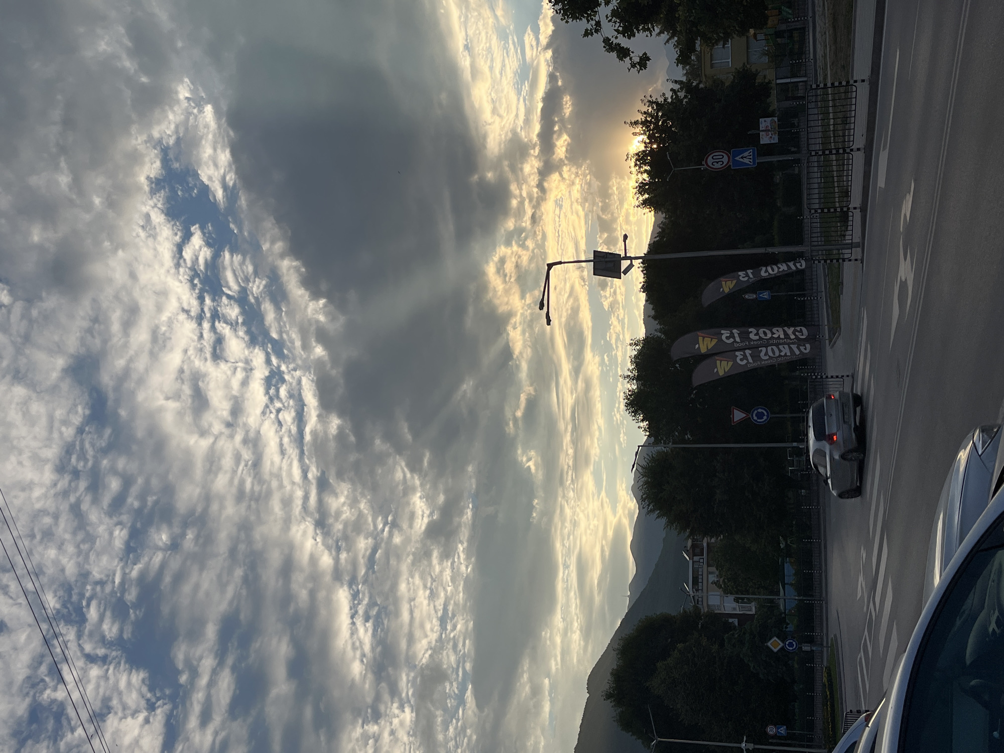 This is a vertical, eye-level shot of a street scene in Gotse Delchev, Bulgaria, bathed in the warm light of a sunset. The sky is filled with dramatic clouds, with rays of sunlight piercing through, creating a sense of awe and serenity.
In the foreground, a grey car is seen driving away from the camera on a paved road with pedestrian crossings. To the right, the front of another white car is partially visible, suggesting it might be parked or also in motion. The road is bordered by a sidewalk and a black metal fence. Several dark green trees line the street, some with branches extending into the frame. Beyond the trees, a few buildings are visible, including one with visible windows. In the distance, a mountain range is silhouetted against the sunset-lit sky.
Traffic signs are present, including a circular sign with a red border and a "30" inside, indicating a speed limit, and a pedestrian crossing sign. Two large, dark banners with white text are displayed on poles along the roadside. The text on the banners is in Cyrillic script and appears to be an advertisement for "GYROS" and "Authentic Greek Food". The time of day is clearly sunset, and the weather appears clear with scattered clouds. The overall mood is peaceful and visually striking, highlighting the beauty of natural light interacting with urban and natural landscapes.