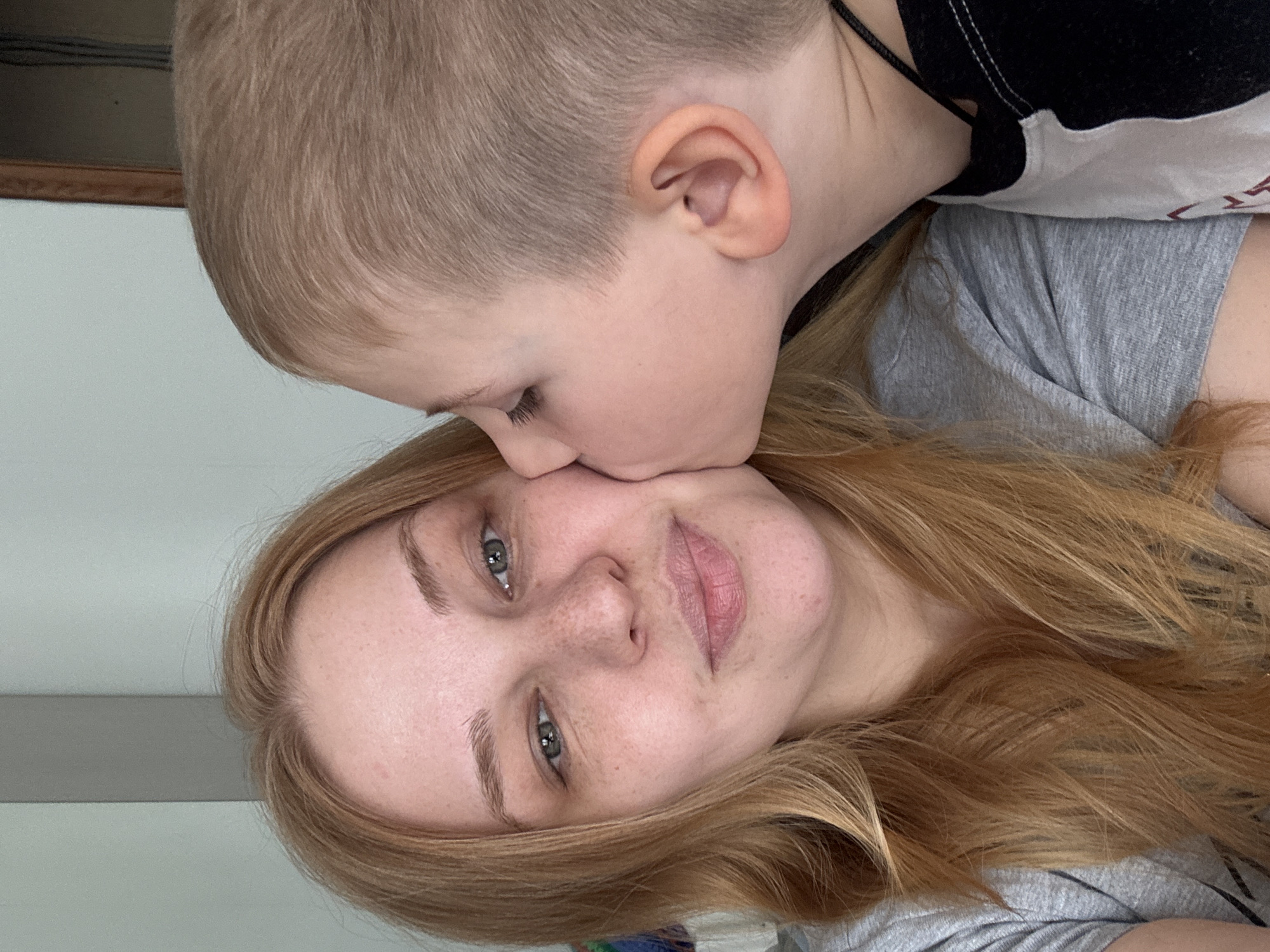 A close-up shot shows a woman and a young child. The woman lies down, facing the camera, with reddish-blonde hair. She has light skin, freckles, and light eyes. A young child with blond hair leans down to kiss her cheek. The child is wearing a black and white top. The woman appears to be wearing a gray shirt. The background includes a section of white wall and a strip of what looks like wood trim. The photograph could have been taken inside a home in Rusanovo, Russia.
