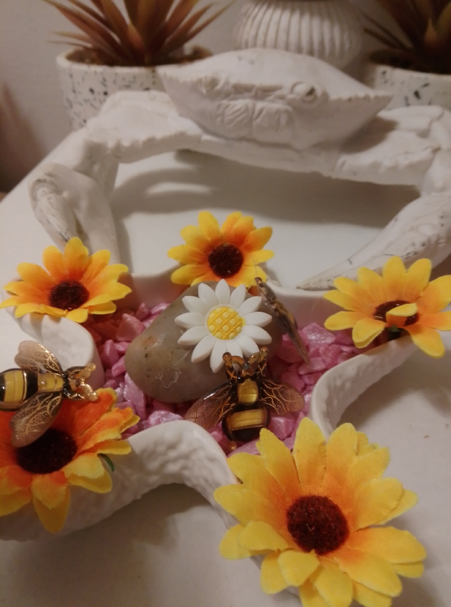 The image depicts a decorative arrangement featuring artificial sunflowers, bumblebee figurines, and a single white daisy on a bed of pink decorative stones. The arrangement is placed on a white, crab-shaped sculpture, which is itself on a white surface. In the background, out of focus, are potted plants and a ribbed ceramic vase. The composition suggests an indoor setting, possibly a home or shop display. There is no indication of the specific location within the city of Wedel, Germany, as no identifying landmarks or contextual clues are visible. The image is a still life, with no people or actions occurring.
