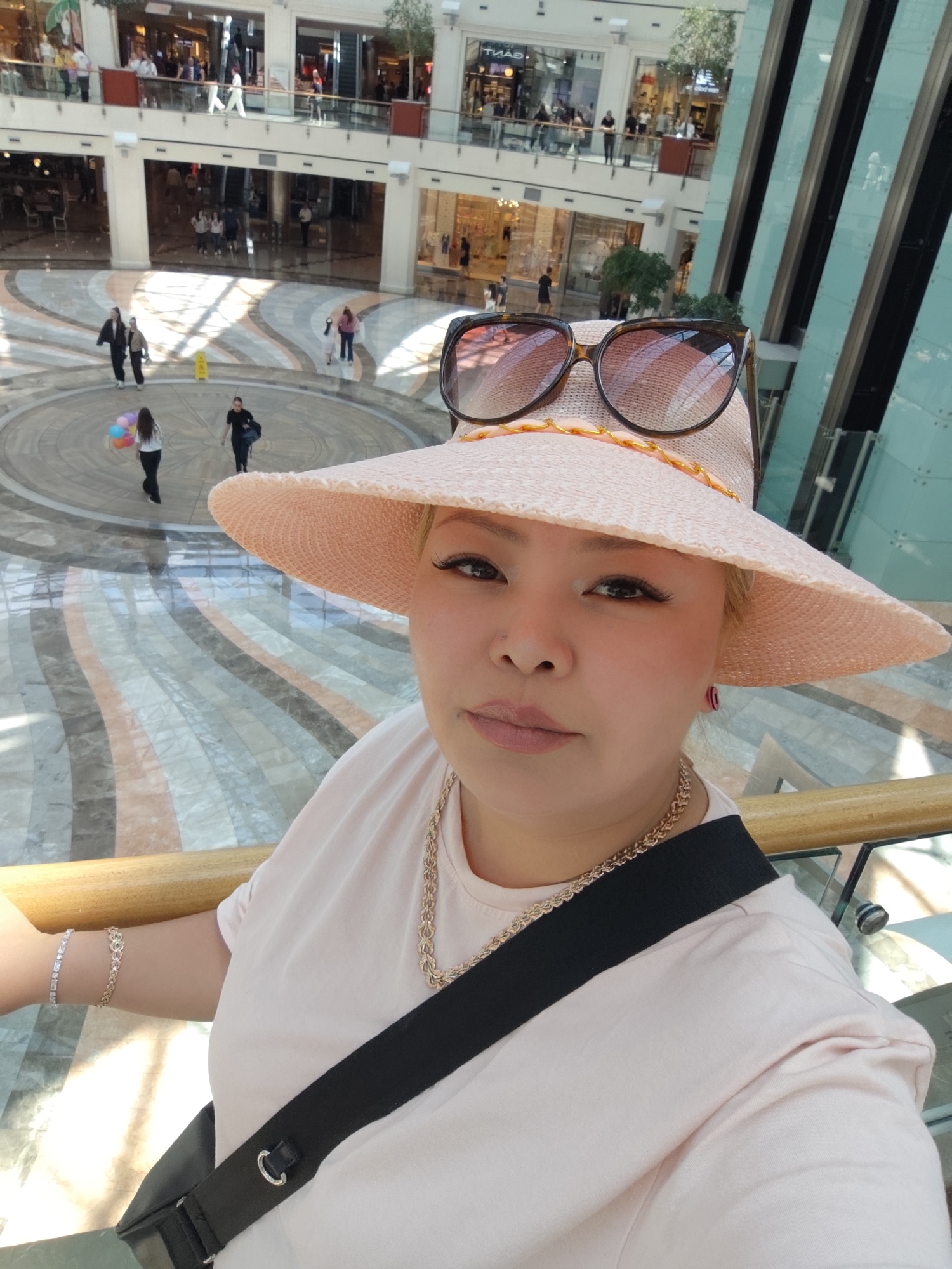 A selfie shows a woman in a large, light pink hat with sunglasses atop, wearing a light pink t-shirt and a gold chain. A black bag strap is slung across her torso. She stands near a wooden railing, overlooking a large indoor space. Below her is a patterned marble floor and other shoppers in the background. Two levels of shops are visible beyond the open space, as are tall panes of glass. The location is a shopping center in Sarıyer, Turkey.