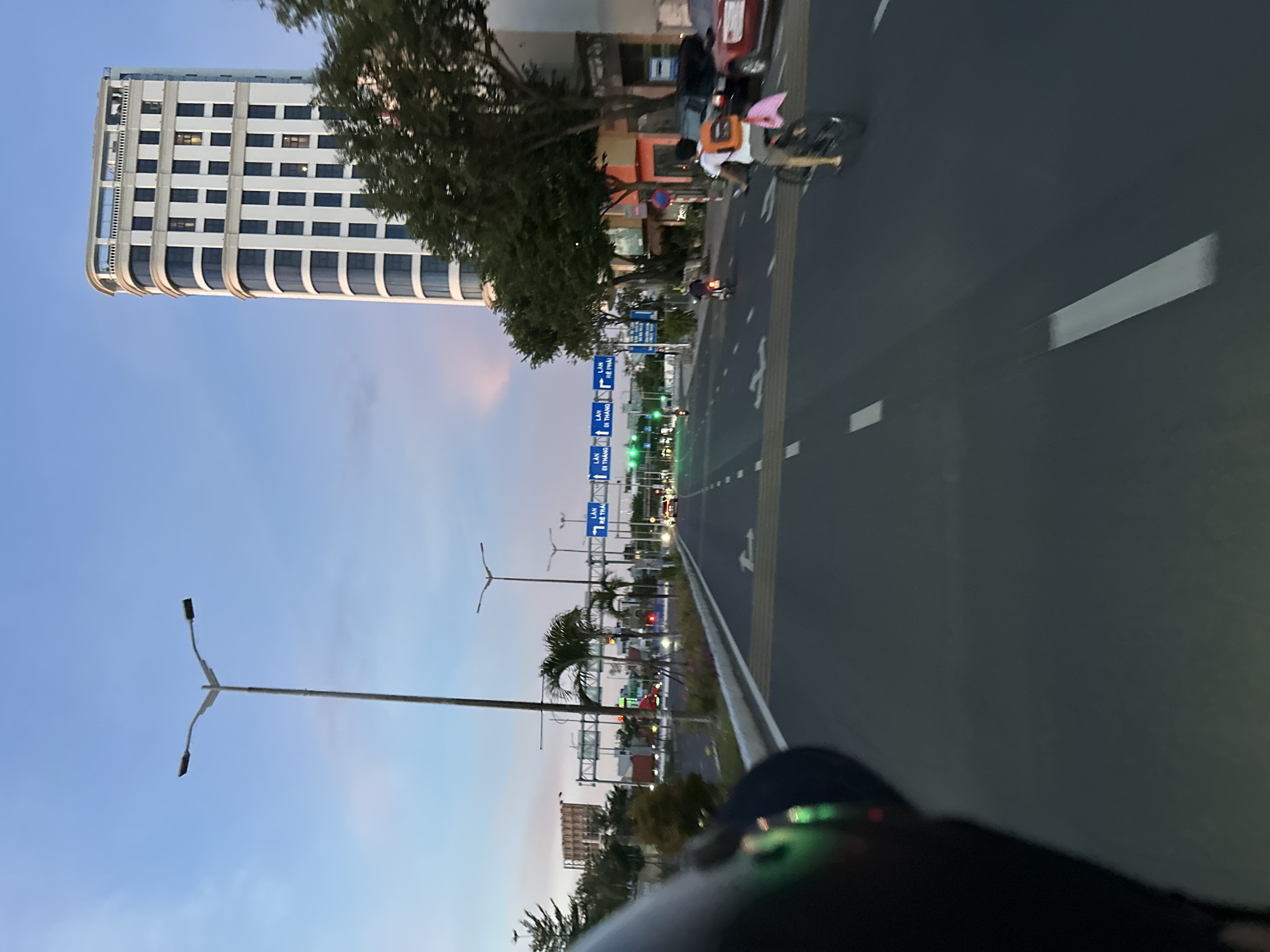 The image shows a wide asphalt road with multiple lanes, indicating a major thoroughfare in a city. A person on a bicycle is visible in the right lane, wearing a bright orange backpack. In the background, there is a tall, modern white building with many windows. Streetlights line the road, and traffic signs with Vietnamese text are also present. The sky is a clear blue with hints of pink and orange, suggesting either sunrise or sunset. Trees and other buildings are scattered along the sides of the road. The perspective is from someone riding a motorcycle, as a portion of a black helmet is visible in the lower left corner. The scene appears to be in Da Nang, Vietnam, based on the presence of Vietnamese text on the signs.