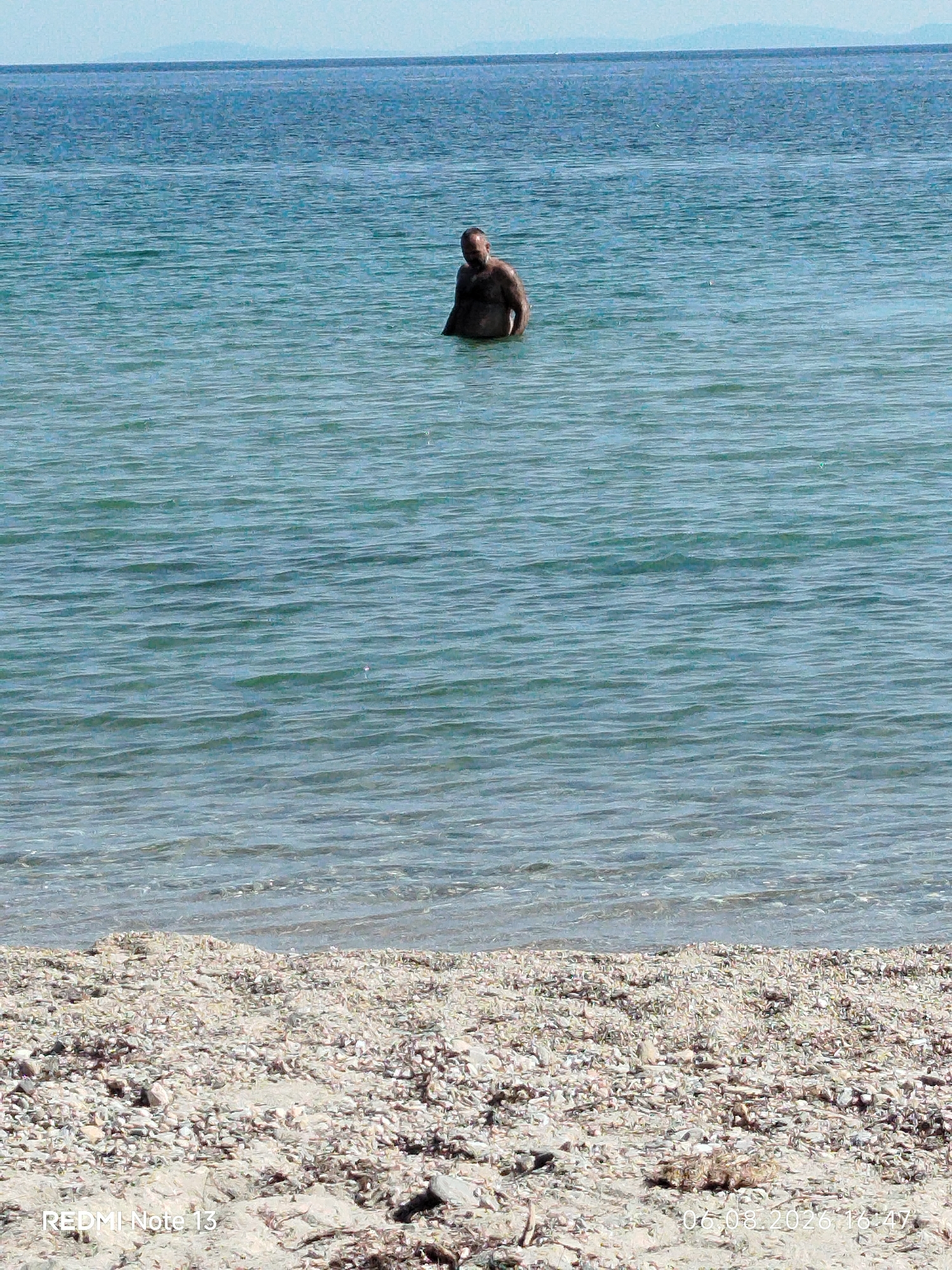 A man stands in the shallow, blue-green water of a beach, with his torso and head visible. The water is calm with subtle ripples. In the background, the horizon shows a faint, hazy line of land or mountains against a pale blue sky. The foreground is a beach covered in small, light-colored pebbles and shells, with some dry vegetation. The overall lighting suggests a bright, sunny day. The timestamp "06.08.2026 16:47" and "REDMI Note 13" are visible in the lower portion of the image. The location is the Sithonia Municipal Unit, Greece, likely a coastal area.