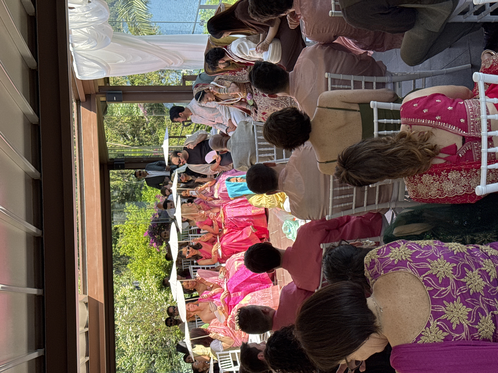 The image shows an outdoor wedding ceremony in Castel Gandolfo, Italy. The ceremony is being held under a white draped structure, possibly a tent or pergola. Guests are seated in rows of white chairs. Many guests are wearing colorful South Asian clothing, including sarees in shades of pink, purple, and gold. Several guests are holding white parasols to shield themselves from the sun. The groom, dressed in white and gold traditional attire, is seen standing with the bride in the aisle. Other people are standing near the entrance, looking at the ceremony. The environment includes trees, greenery, and possibly a lake in the background, seen through the structure. A building with windows is on the left side of the image.