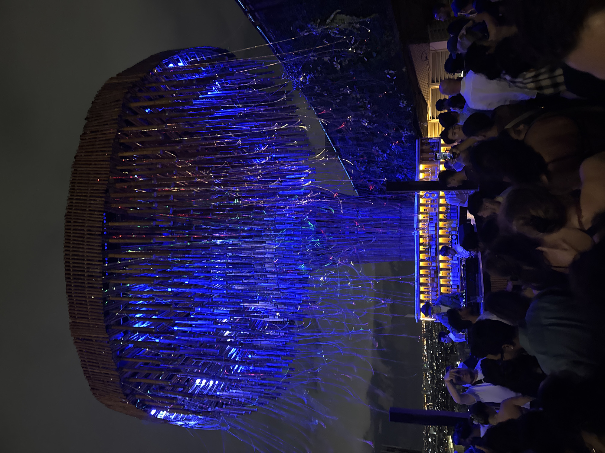 This image captures a bustling nighttime scene in Bangkok, Thailand, likely at a rooftop bar or event space. The photograph has been taken at an angle and then rotated.
On the left is a large, basket-like structure or sculpture, possibly made of bamboo or similar natural materials, that is illuminated with bright blue lights. The blue illumination casts shadows and provides the main light source for this area.
On the right of the structure, a crowd of people is gathered. Some are turned toward a brightly lit bar area that is partially visible behind them, showing shelves filled with bottles. In the background, a city skyline stretches out, dotted with lights. The sky is dark and cloudy, indicative of a nighttime or early morning setting. The overall ambiance is vibrant and energetic, suggesting an active social gathering.